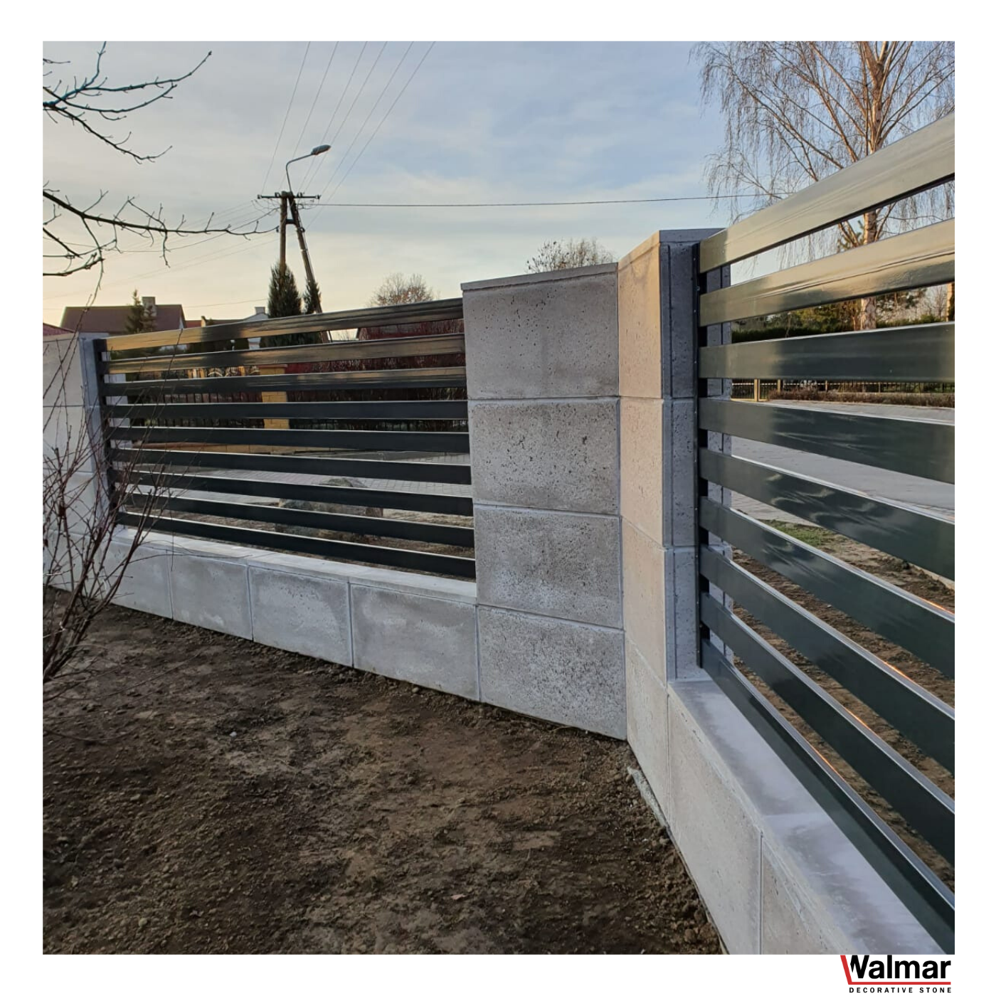 Nowoczesne ogrodzenie panelowe z betonowymi słupkami i podmurówką, poziome przęsła w kolorze grafitowym, widoczny fragment ogrodu i słup energetyczny w tle.