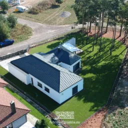 Widok z lotu ptaka na nowoczesny dom z ciemnym dachem, otoczony intensywnie zielonym trawnikiem, graniczącym z brązową korą i wysokimi drzewami rzucającymi długie cienie.