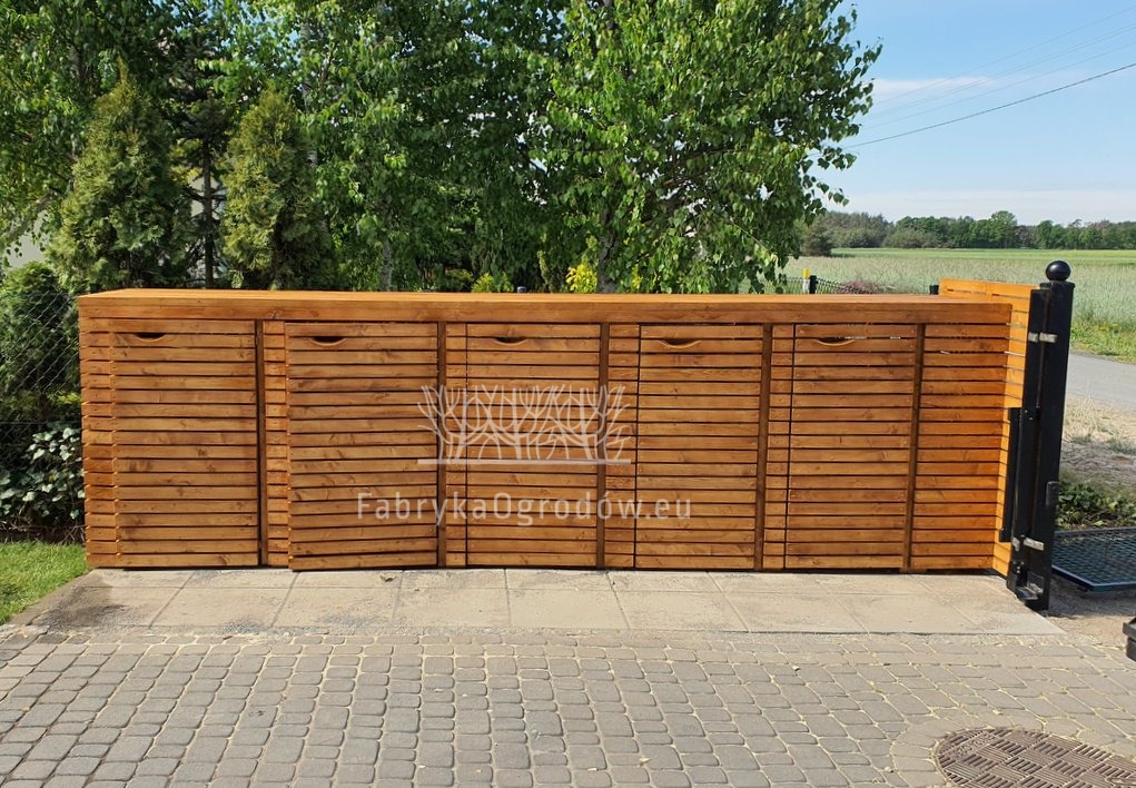 Drewniana zabudowa śmietnikowa z lamelami, zintegrowana z ogrodzeniem, na tle zieleni. Estetyczne rozwiązanie do przechowywania pojemników na odpady.