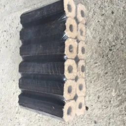 Zapakowany w folię stos brykietu drzewnego z charakterystycznym otworem w środku, leżący na szarej betonowej powierzchni.