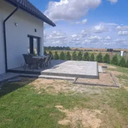 Szara kostka brukowa ułożona na tarasie przylegającym do białego domu, z widocznym stołem i krzesłami. Część terenu przygotowana pod dalsze prace brukarskie, w tle zielona przestrzeń z drzewami...
