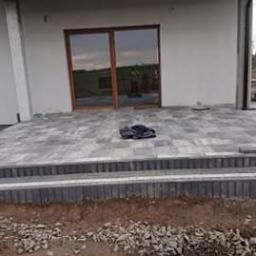 Usługi Brukarskie Shkvarlo - Nowo ułożony taras z kostki brukowej w odcieniach szarości, z dwoma stopniami, przed białym budynkiem z dużymi przesuwnymi drzwiami tarasowymi; widoczny pracownik w roboczych ubraniach i rękawicach.