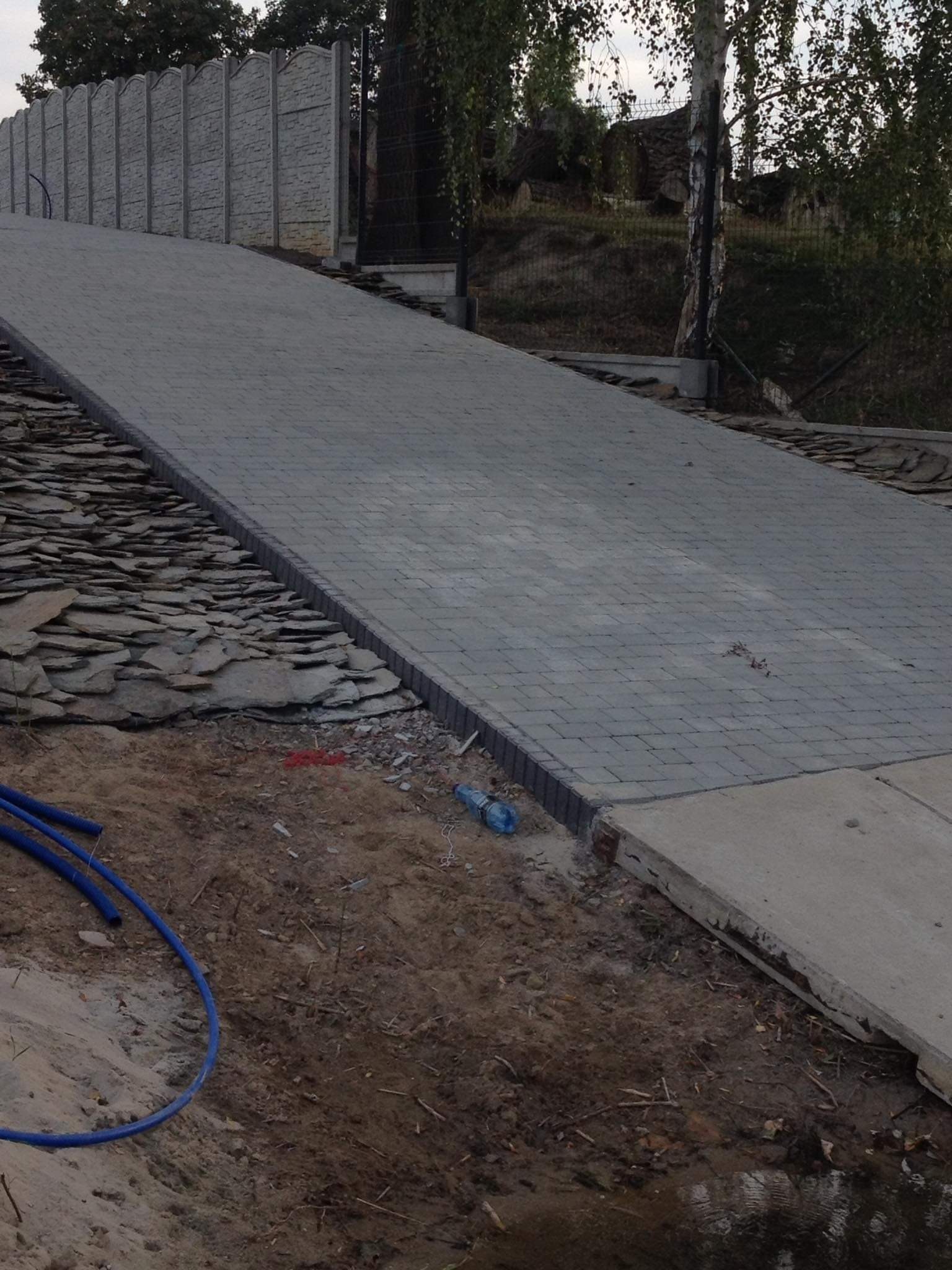 Ukończona ścieżka z szarej kostki brukowej prowadząca pod górę, obrzeża wykończone kamieniem łupkowym, w tle betonowe panele ogrodzeniowe.