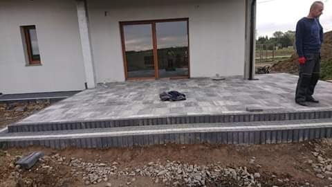 Nowo ułożony taras z kostki brukowej w odcieniach szarości, z dwoma stopniami, przed białym budynkiem z dużymi przesuwnymi drzwiami tarasowymi; widoczny pracownik w roboczych ubraniach i rękawicach.