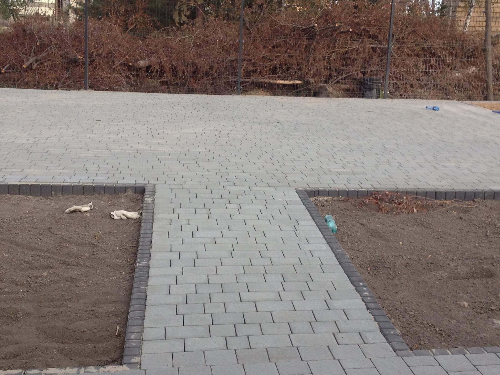 Ułożona szara kostka brukowa z obrzeżami, widoczne dwie rękawice robocze i butelka plastikowa, w tle gałęzie za ogrodzeniem.