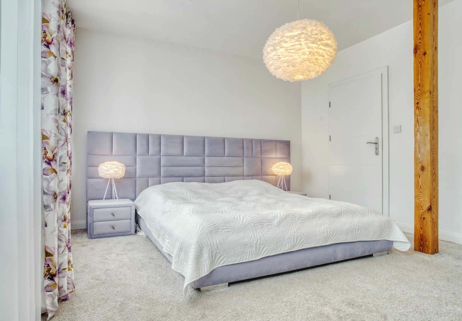 Jasna sypialnia z łóżkiem z szarym tapicerowanym zagłówkiem, puszystą lampą wiszącą i drewnianą kolumną obok białych drzwi.