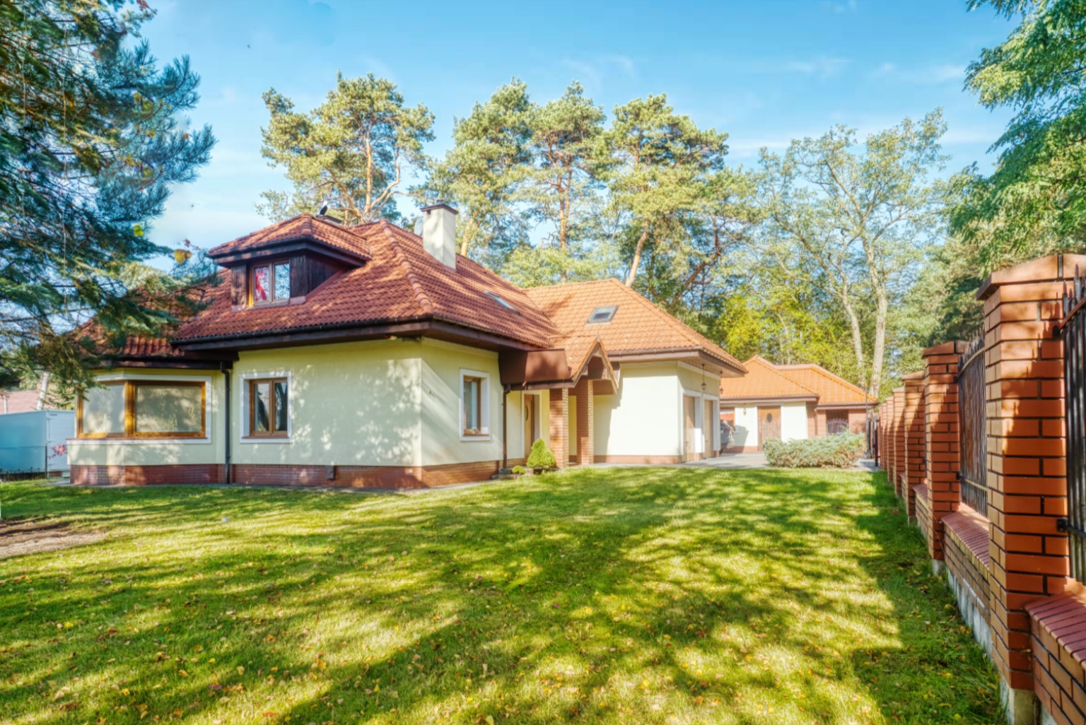 Jasny, parterowy dom z czerwonym dachem, otoczony zielonym trawnikiem i drzewami, widoczny zza ceglanego ogrodzenia z metalowymi przęsłami.