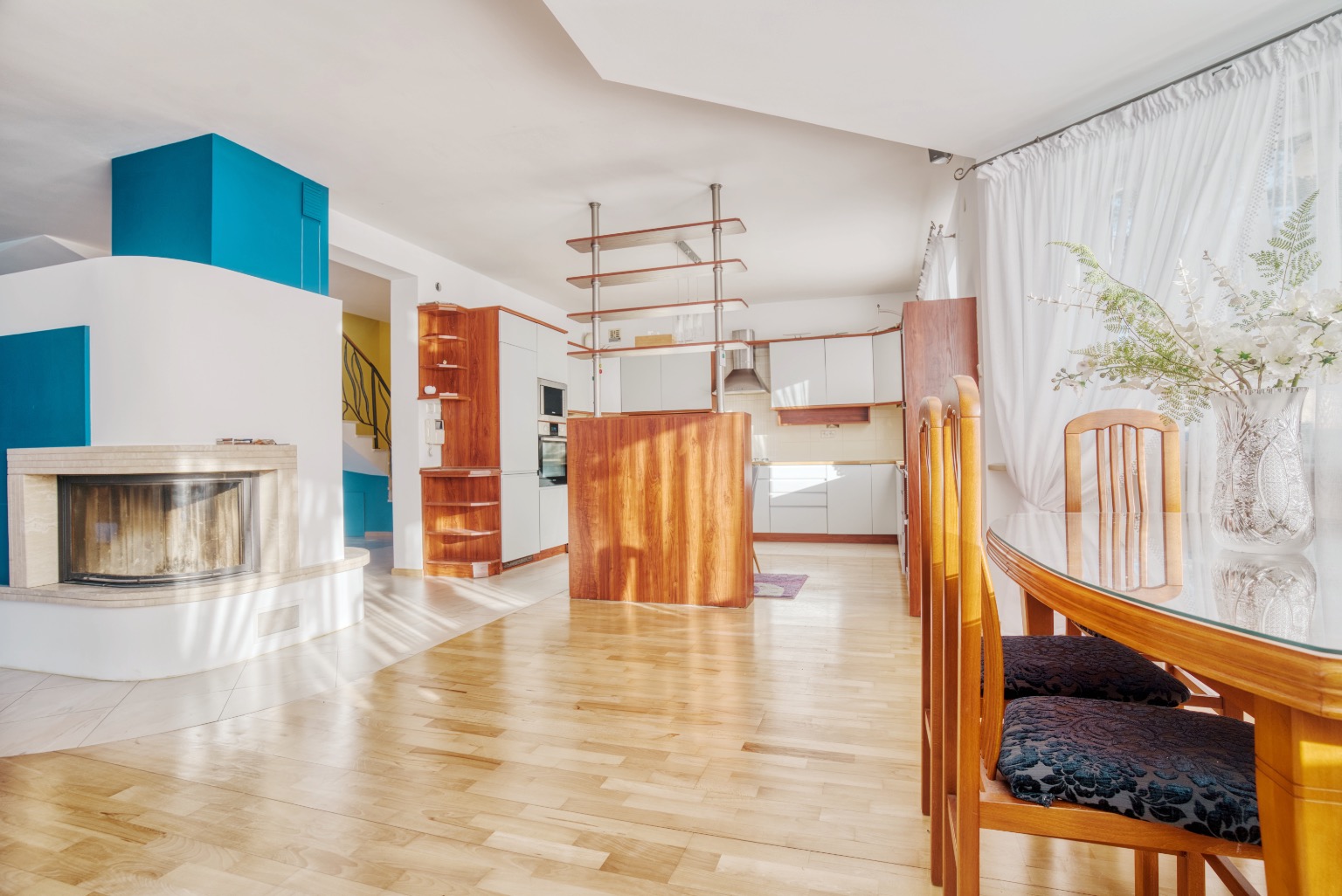 Jasne, przestronne wnętrze: parkiet, kominek z turkusowym akcentem, kuchnia z drewnianą wyspą i stół z kwiatami w wazonie.