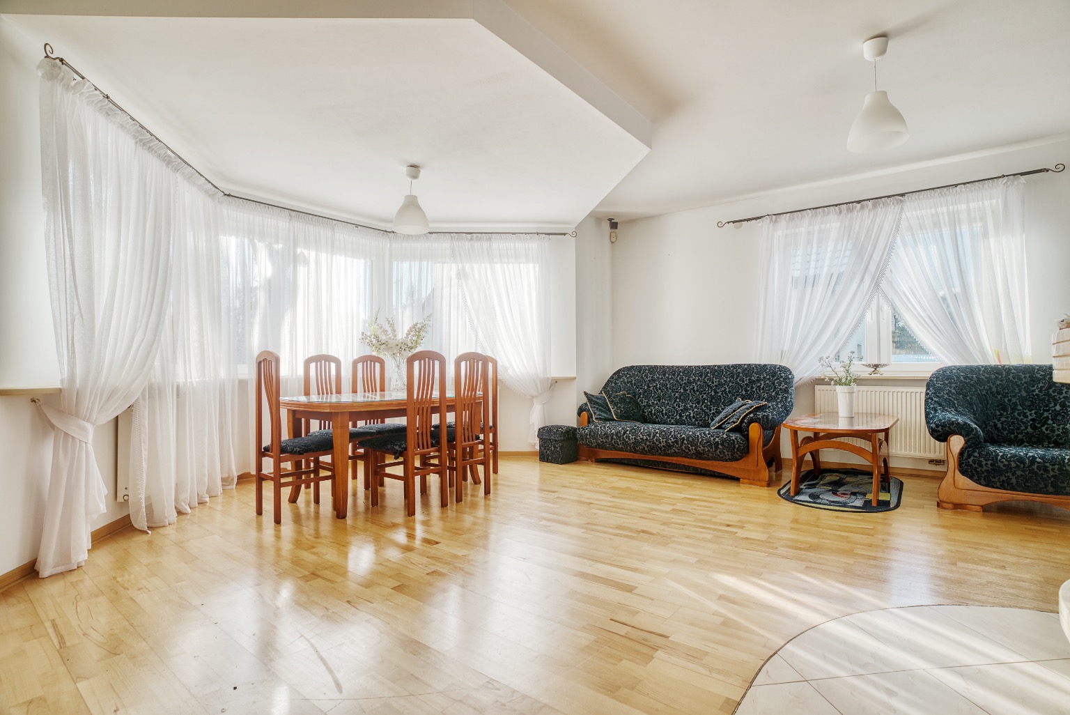 Jasne wnętrze salonu z drewnianą podłogą, stołem jadalnym z sześcioma krzesłami, ciemną sofą i fotelem w kwiatowy wzór, okna zasłonięte białymi firanami.