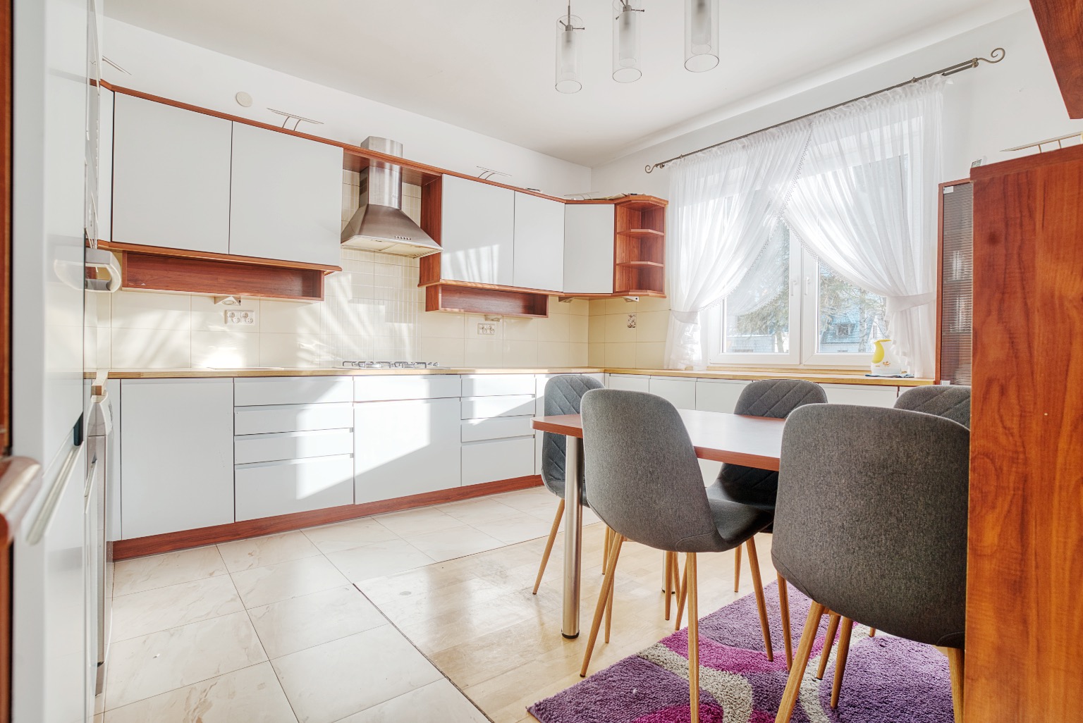 Jasna kuchnia z białymi szafkami, drewnianymi akcentami i szarymi krzesłami wokół stołu, oświetlona naturalnym światłem wpadającym przez okno z firankami.