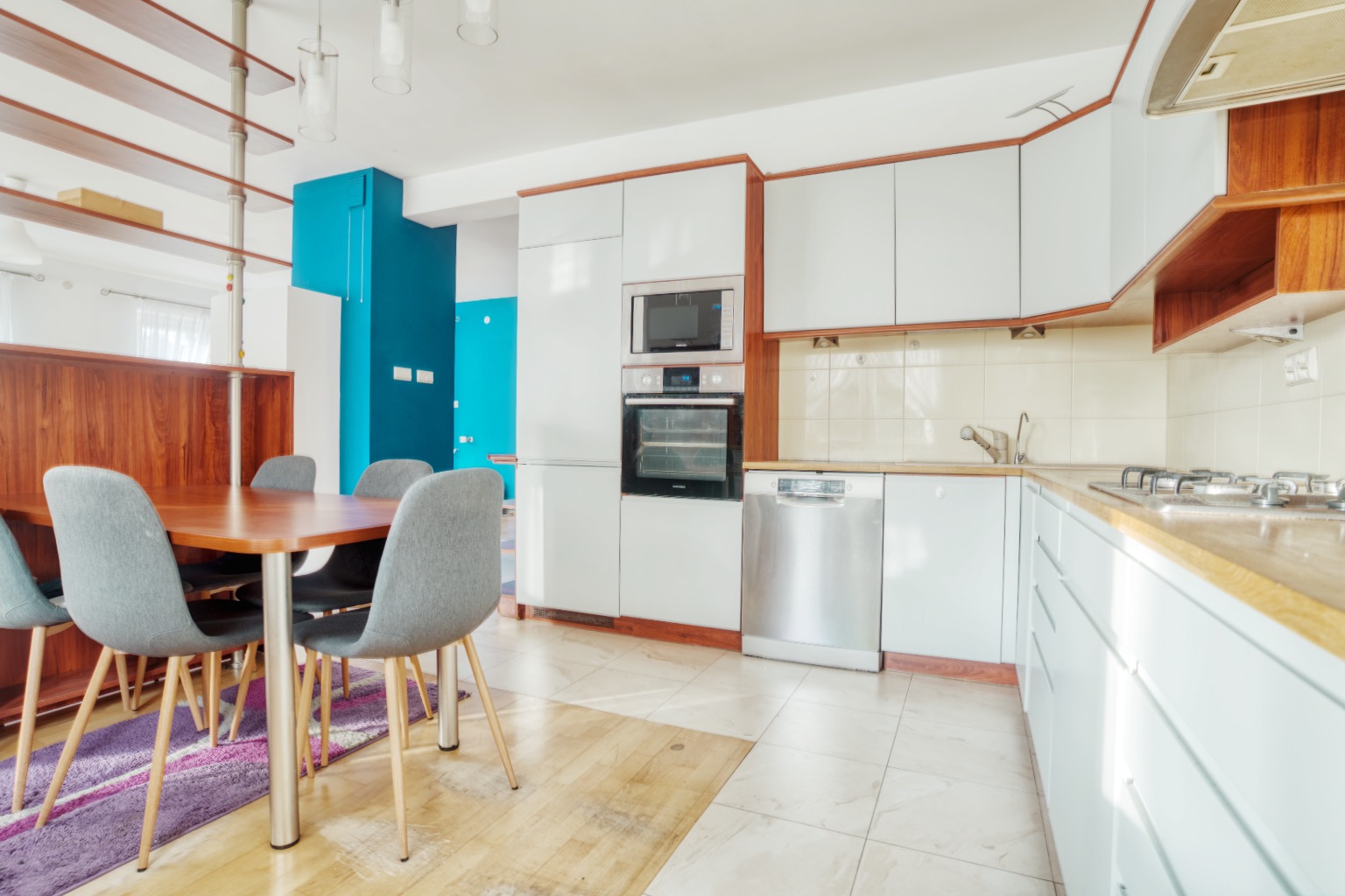 Jasna kuchnia z drewnianymi akcentami, białymi szafkami i turkusową ścianą w tle, stół z szarymi krzesłami na pierwszym planie.