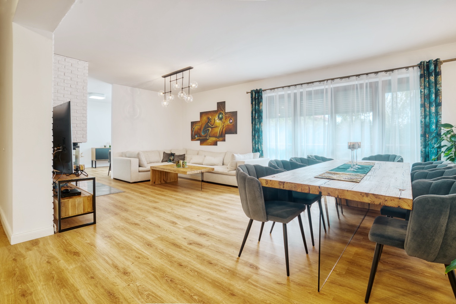 Jasne, przestronne wnętrze salonu z jadalnią, drewniany stół z szarymi krzesłami, biała sofa narożna, drewniany stolik kawowy i dekoracje ścienne.