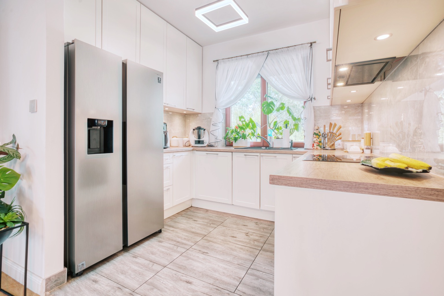 Jasna, przestronna kuchnia w Otwocku z nowoczesną zabudową, dużym srebrnym dwudrzwiowym lodówko-zamrażarką z dystrybutorem wody, drewnianymi blatami i oknem z białymi firankami, doświetlona...