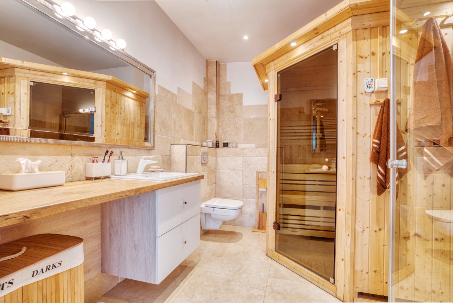 Jasna łazienka z drewnianą sauną i beżowymi płytkami, widoczny blat z umywalką, lustro z oświetleniem, wisząca toaleta i kosz na pranie z napisem 'DARKS'.