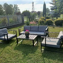 Zestaw mebli ogrodowych z metalową ramą i szarymi poduszkami na trawniku, z kwiatami w doniczce na stoliku kawowym, na tle ogrodzenia i zieleni.