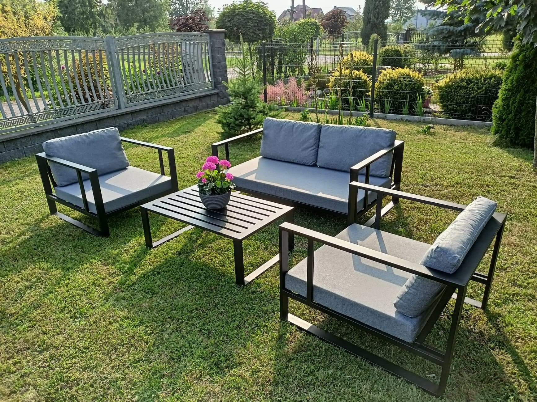 Zestaw mebli ogrodowych z czarną ramą i szarymi poduszkami na trawniku, z kwiatami w doniczce na stoliku, w tle ogrodzenie i zieleń.