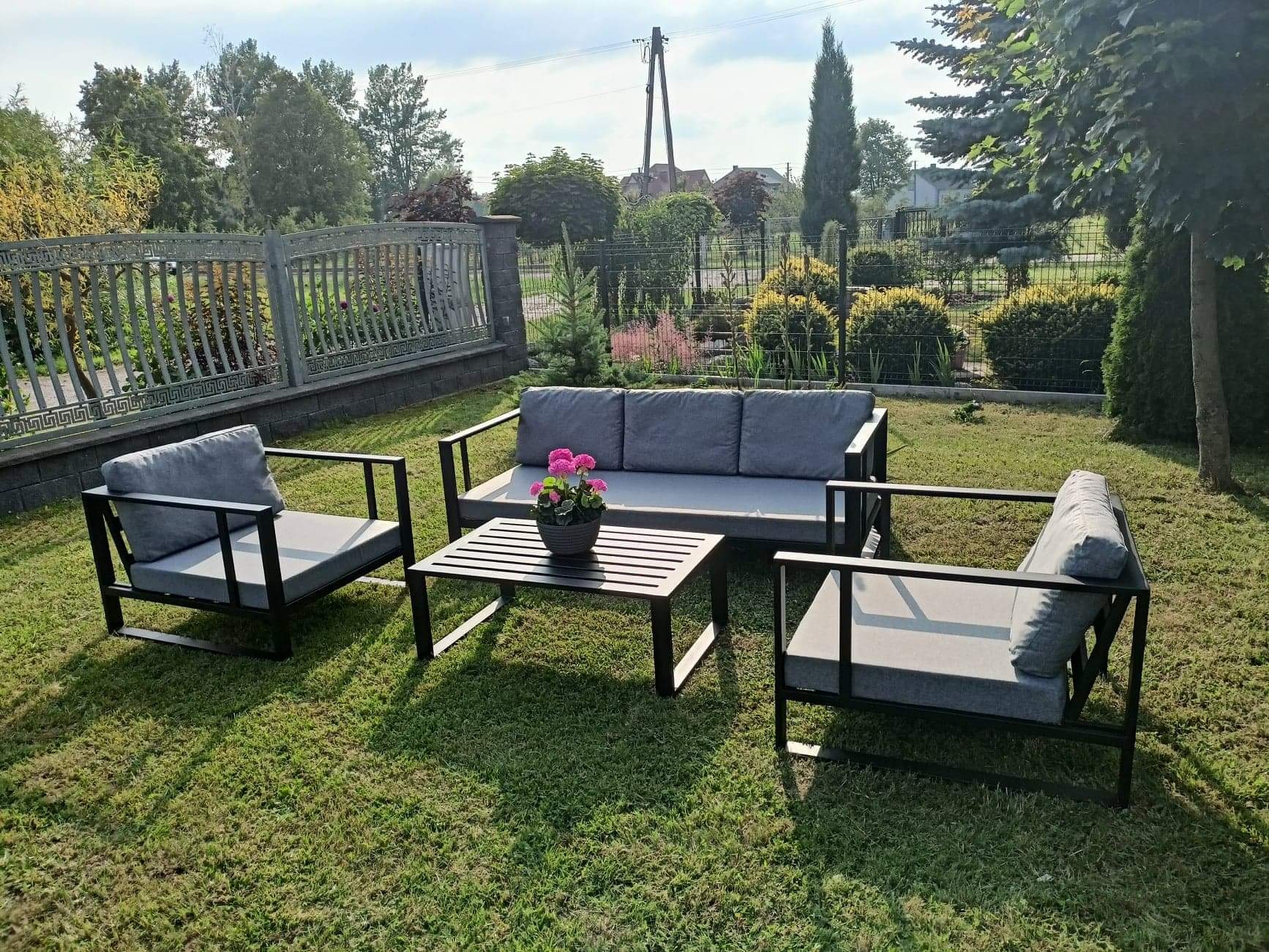 Zestaw mebli ogrodowych z metalową ramą i szarymi poduszkami na trawniku, z kwiatami w doniczce na stoliku kawowym, na tle ogrodzenia i zieleni.