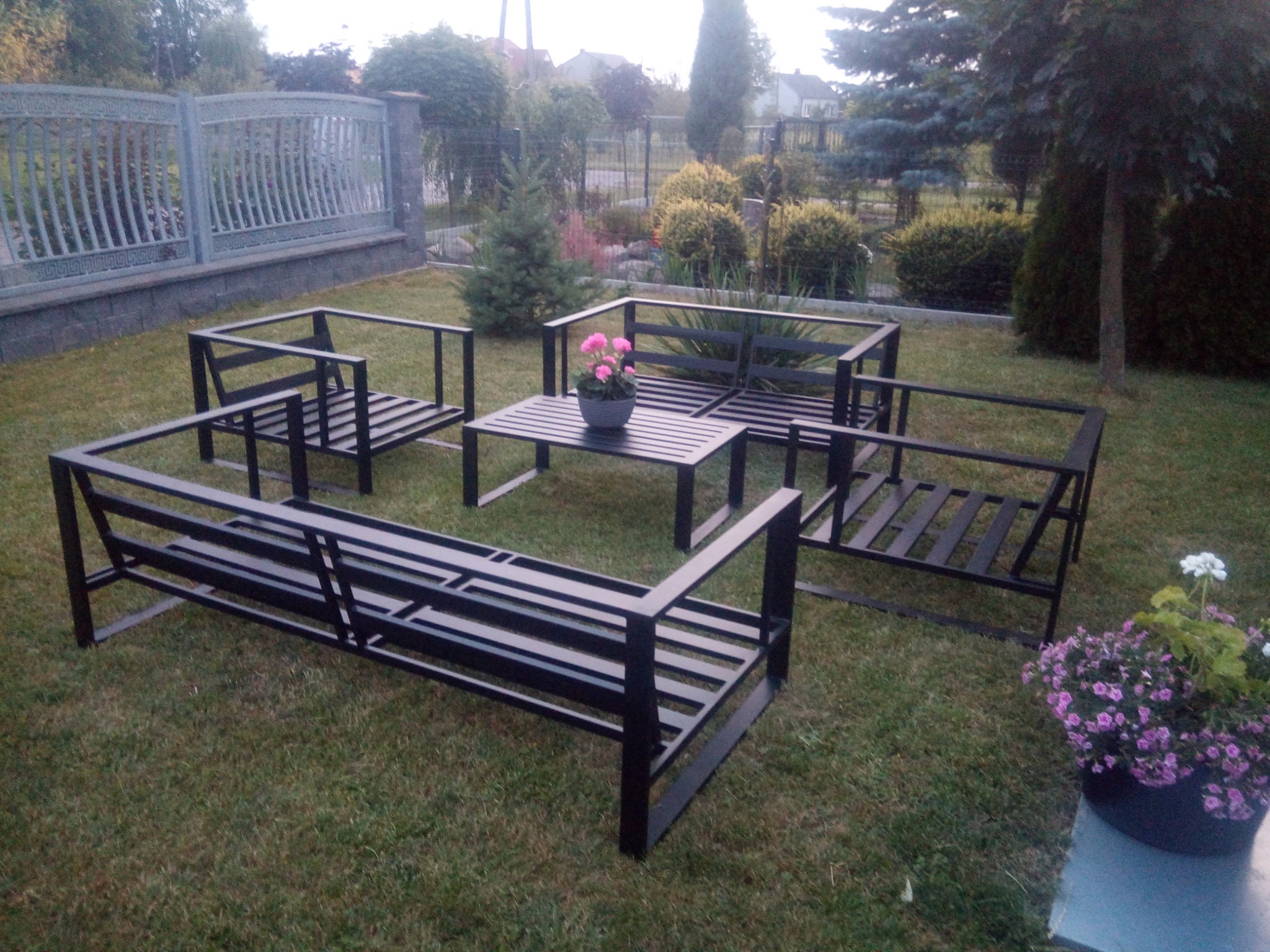 Metalowy zestaw mebli ogrodowych w stylu industrialnym, składający się z dwóch sof, dwóch krzeseł i stolika kawowego, ustawiony na trawniku w ogrodzie z ozdobnymi krzewami i drzewami w tle.