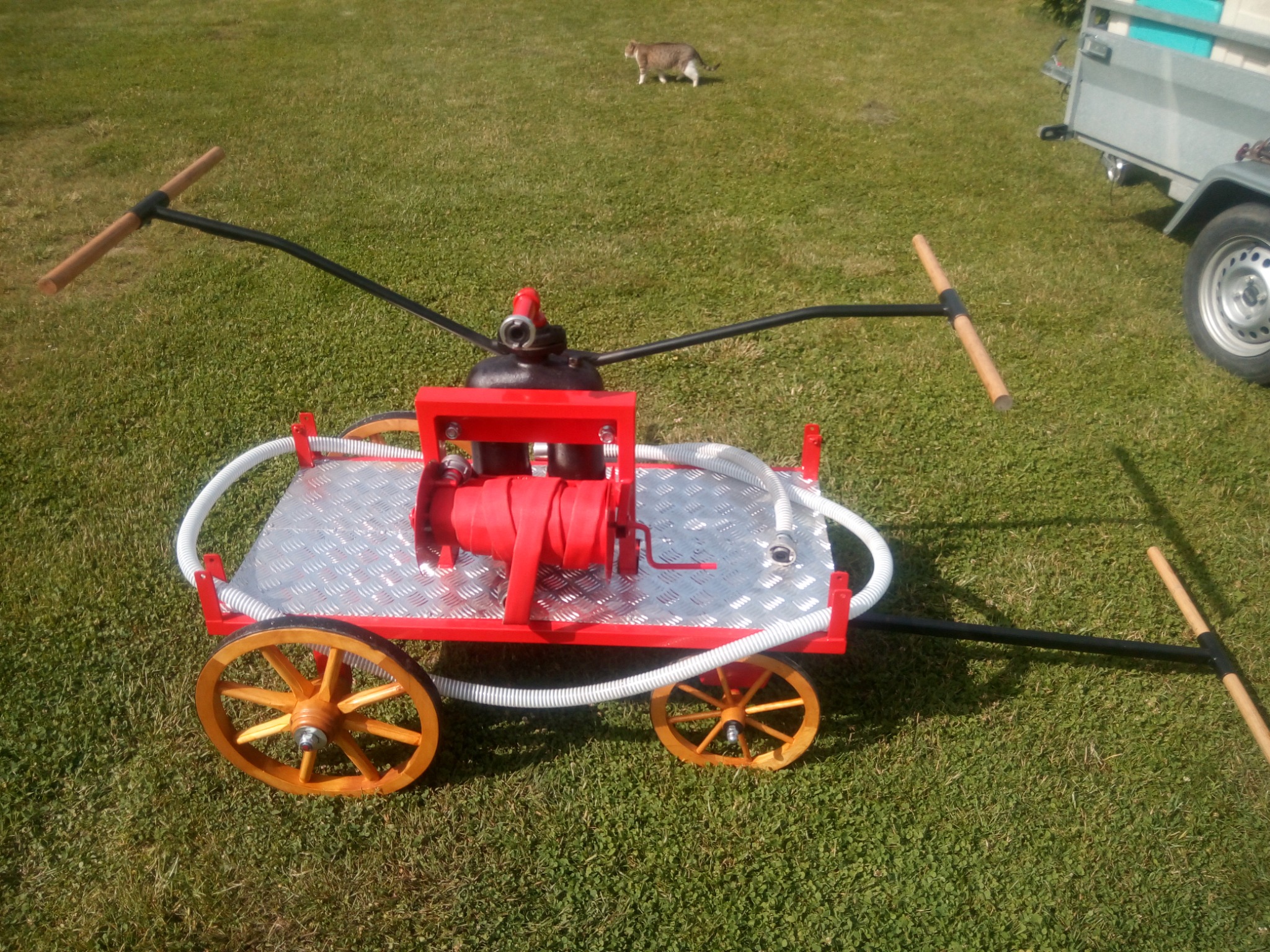 Czerwono-srebrny wózek z drewnianymi kołami, prawdopodobnie przenośna pompa, na trawniku z widocznym kotem i przyczepką samochodową w tle.