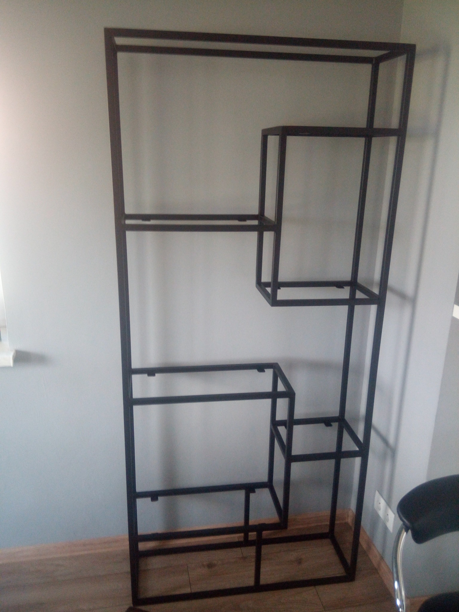 Metalowa konstrukcja regału o asymetrycznym układzie półek, spawana z profili, w surowym stylu industrialnym, na tle szarej ściany i drewnopodobnej podłogi.