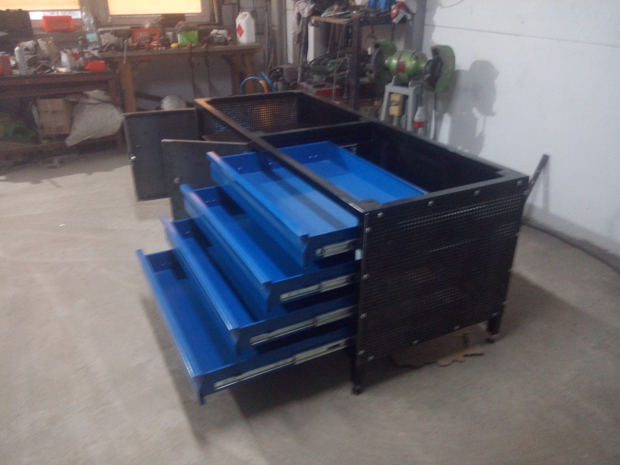 Czarna, metalowa szafka warsztatowa z wysuwanymi, niebieskimi szufladami, widoczna w warsztacie z narzędziami w tle.