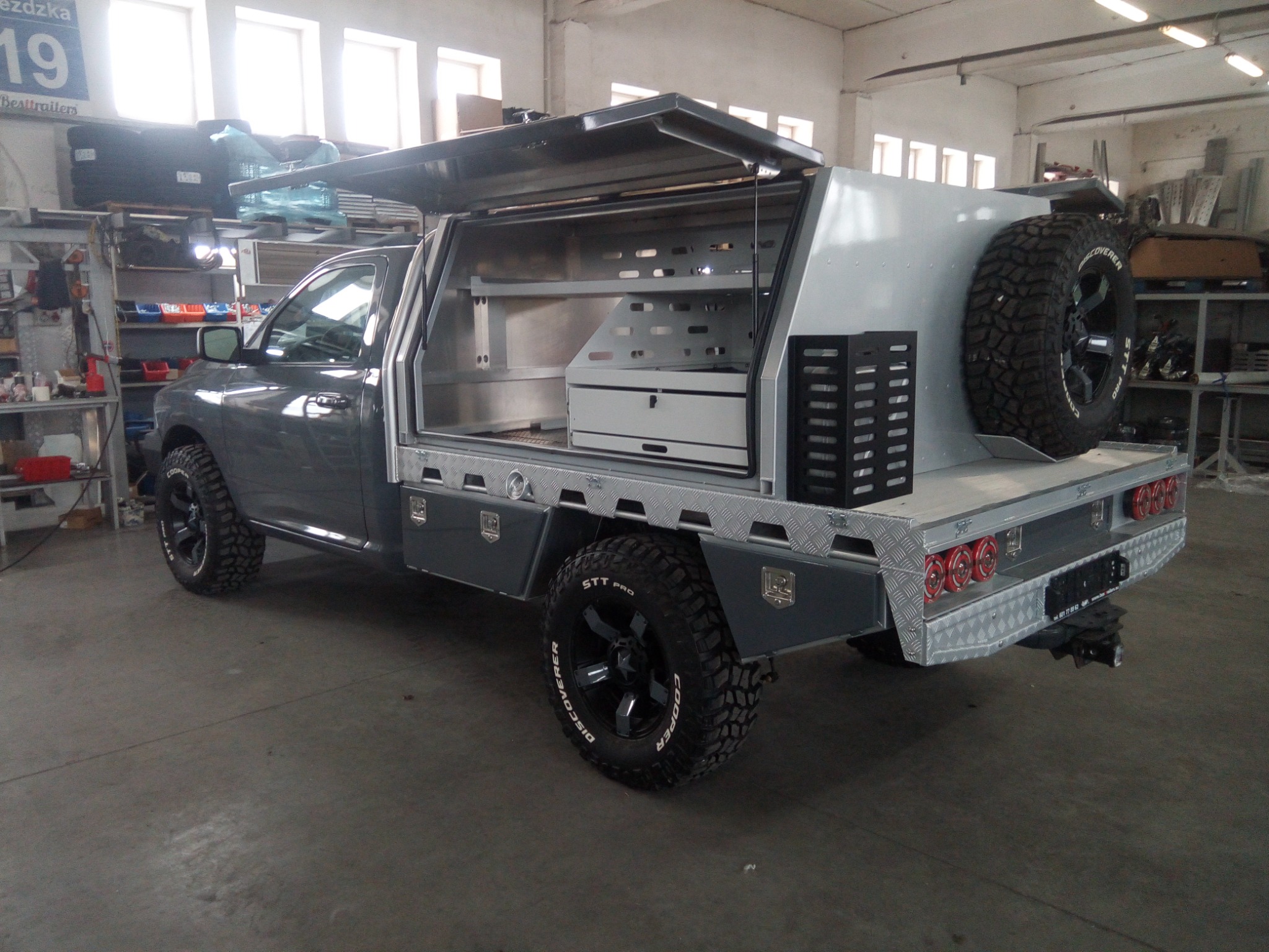 Szara terenówka z zabudową warsztatową na pace, otwartą do góry, z dodatkowym kołem zapasowym, widok z tyłu i boku w warsztacie.