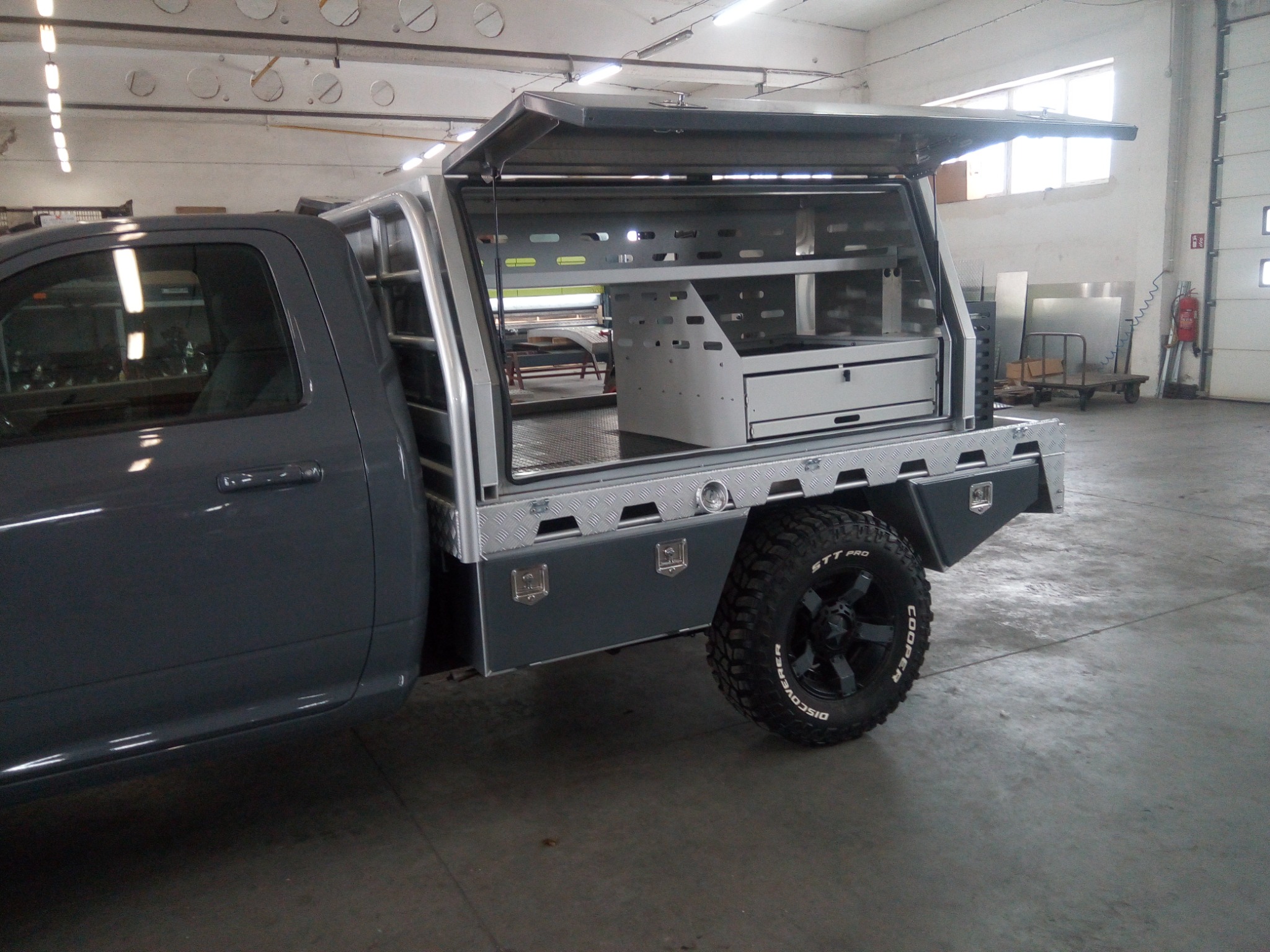 Szary pickup z aluminiową zabudową warsztatową na pace, otwartą do góry, prezentującą wewnętrzne półki i szuflady, z widocznymi oponami terenowymi.