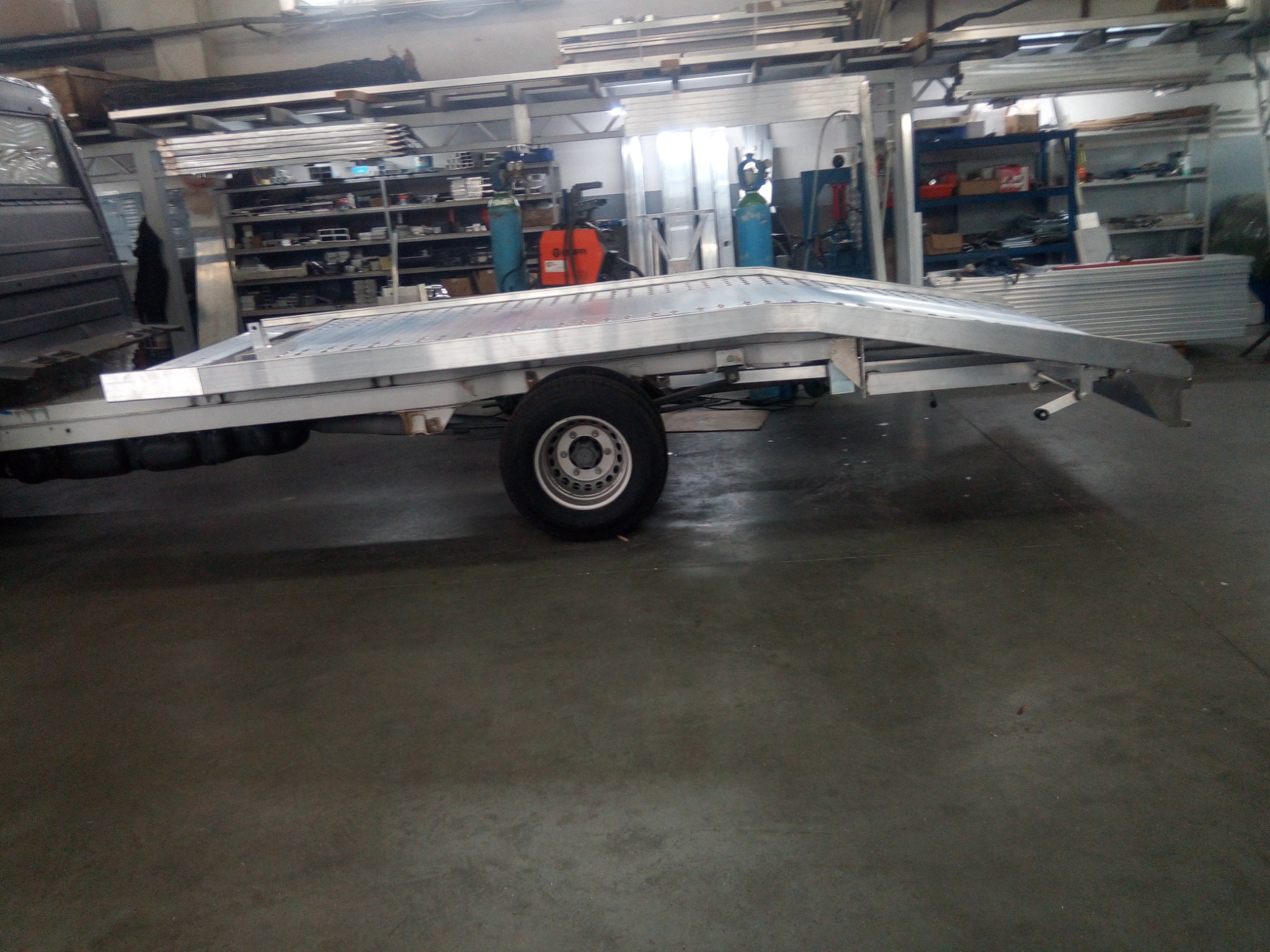 Aluminiowa laweta samochodowa z pojedynczą osią, przymocowana do pojazdu, widoczna w warsztacie z narzędziami i materiałami na półkach w tle.