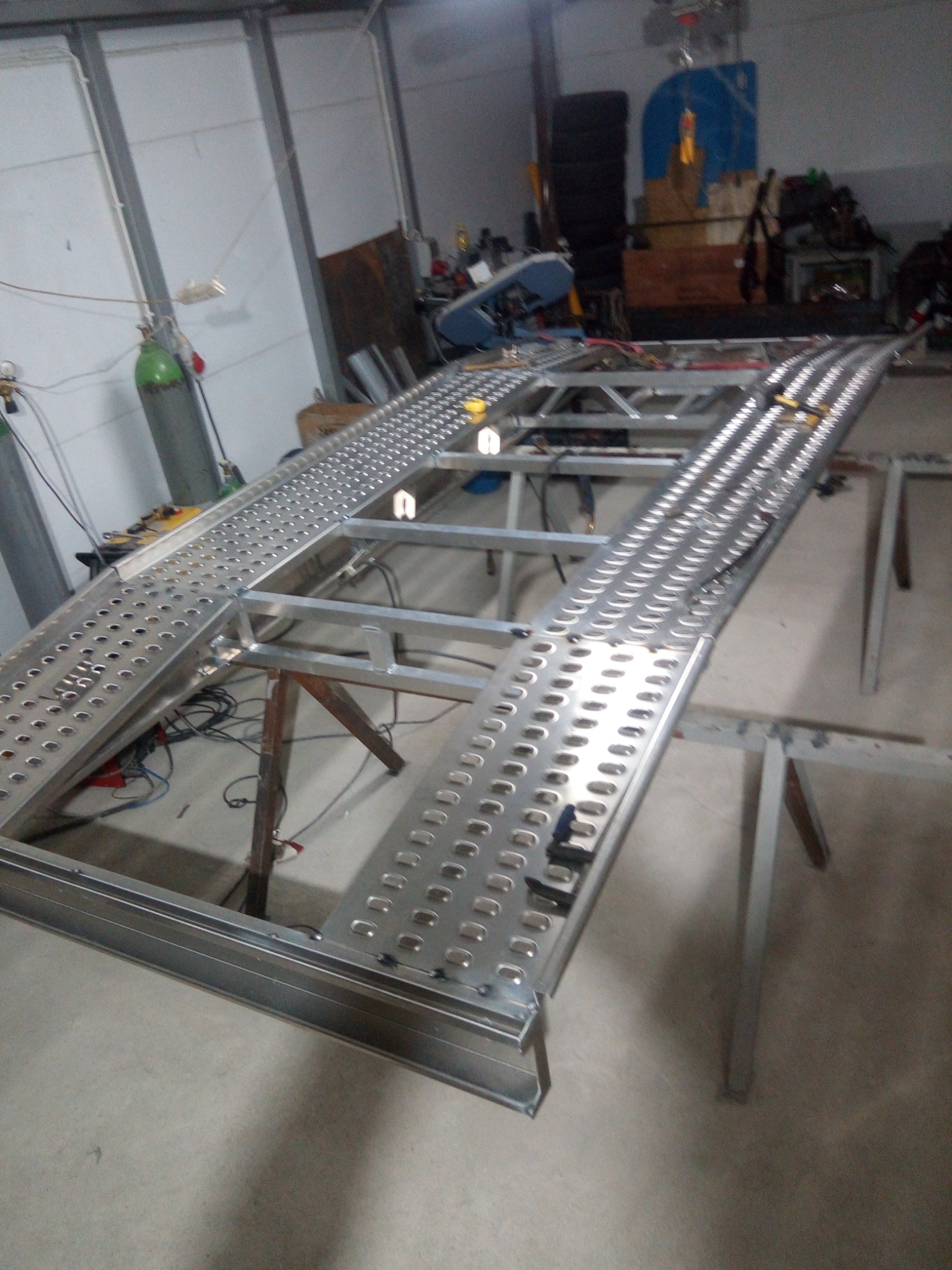 Metalowa rampa najazdowa w trakcie budowy, widoczne spawane elementy konstrukcyjne i perforowana powierzchnia, surowe oświetlenie warsztatowe.