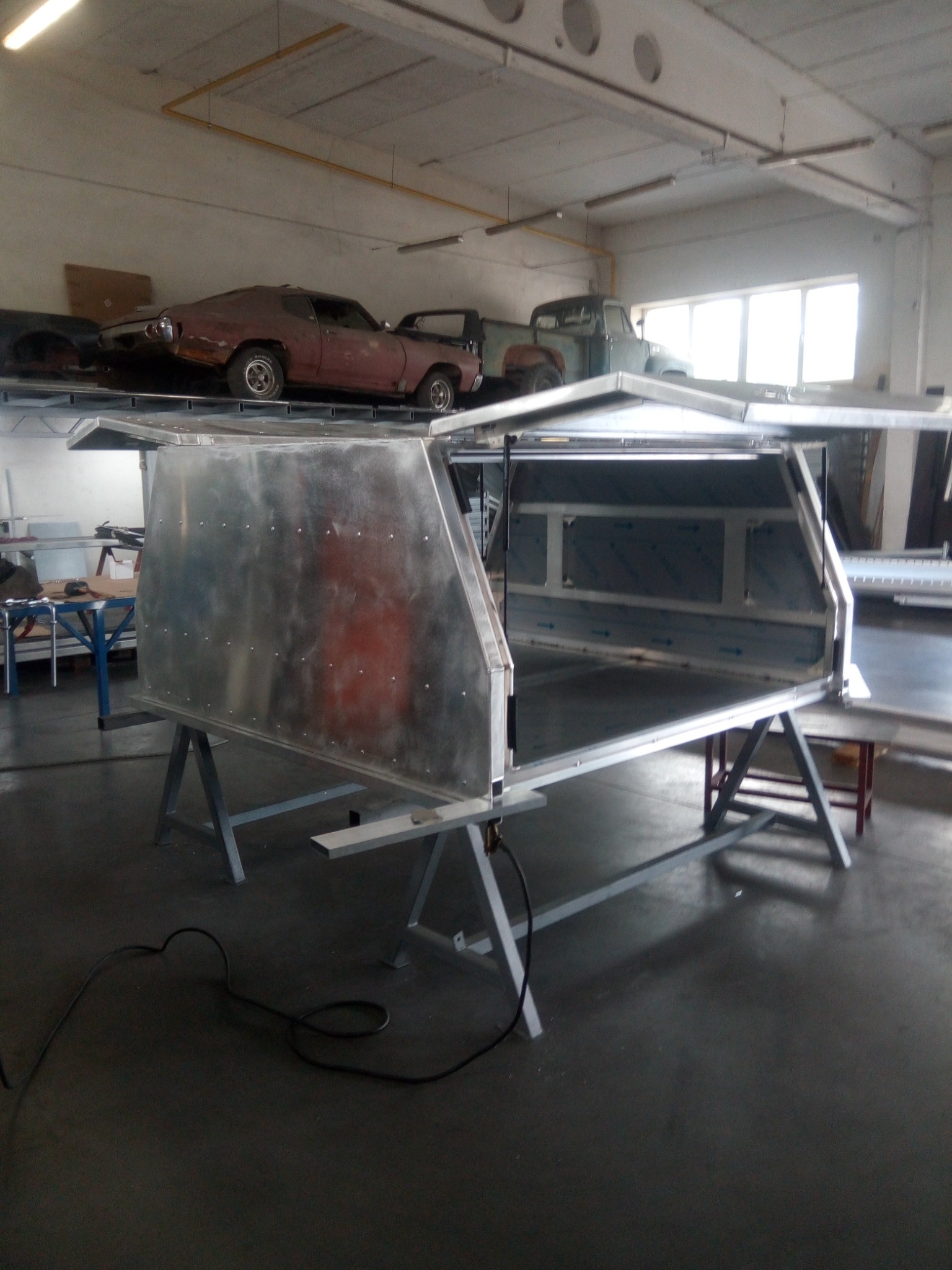 Aluminiowa zabudowa przestrzeni ładunkowej pickupa na metalowych kozłach, w tle zaparkowane klasyczne samochody w warsztacie.