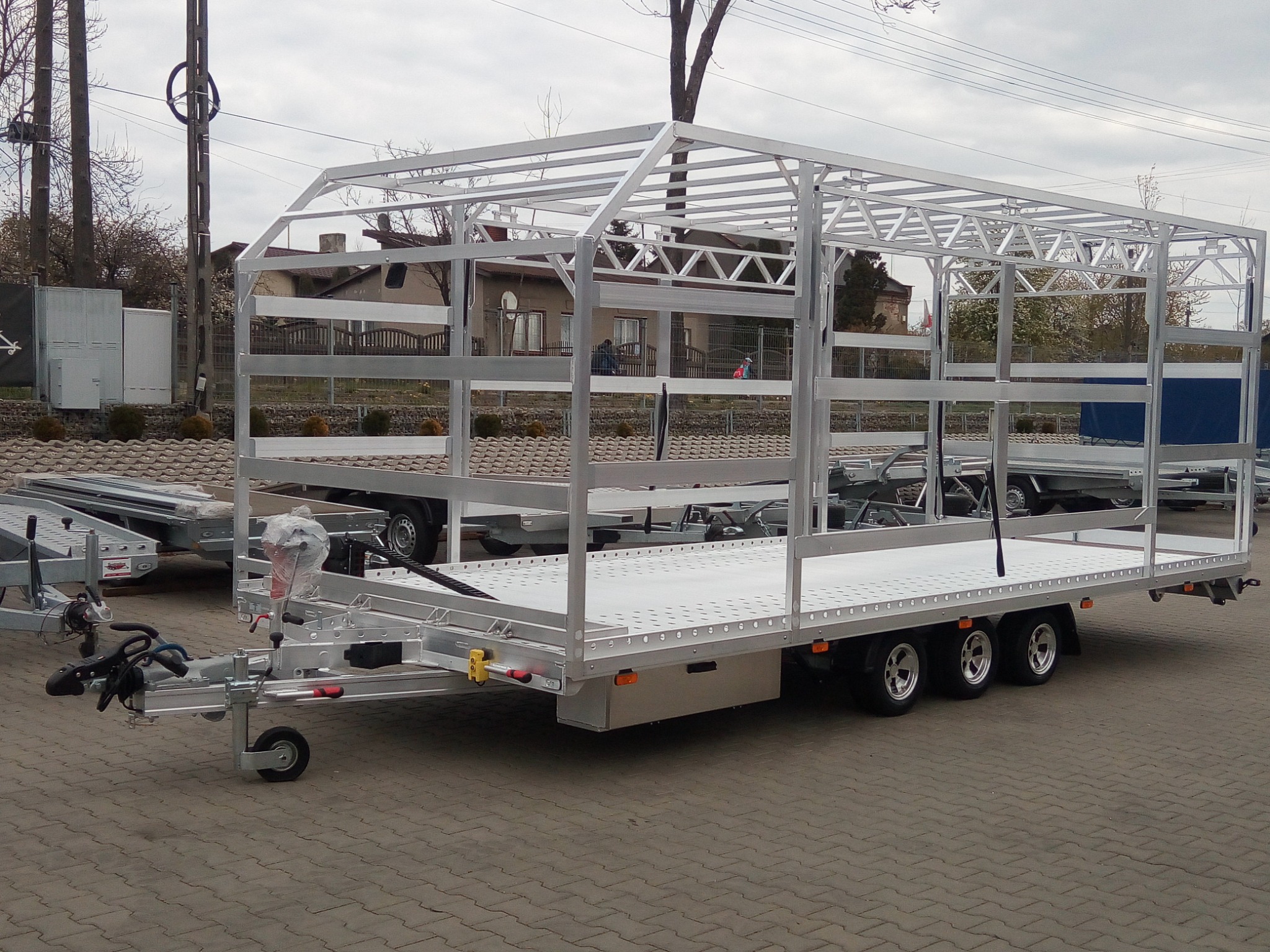 Nowoczesna, aluminiowa przyczepa z ramą i trzema osiami, stojąca na brukowanym placu, z widocznymi detalami konstrukcyjnymi i zabezpieczeniami.