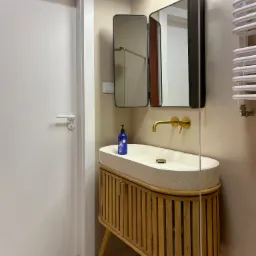 Nowoczesna łazienka z umywalką z terazzo na drewnianej szafce z lamelami, złotą armaturą i lustrem z otwieranymi skrzydłami.