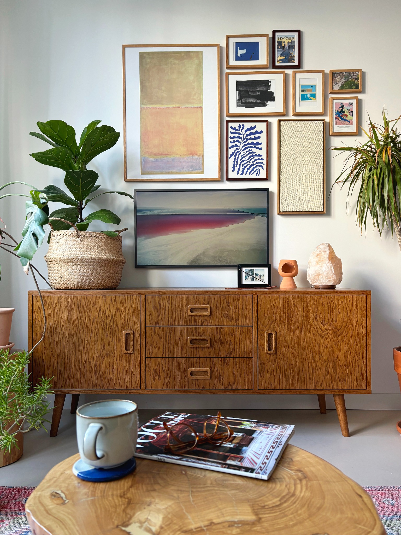 Salon w stylu retro z drewnianym kredensem, na którym stoi telewizor, lampa solna i dekoracje. Na ścianie galeria obrazów i rośliny doniczkowe. Na stoliku kawa, okulary i magazyn.