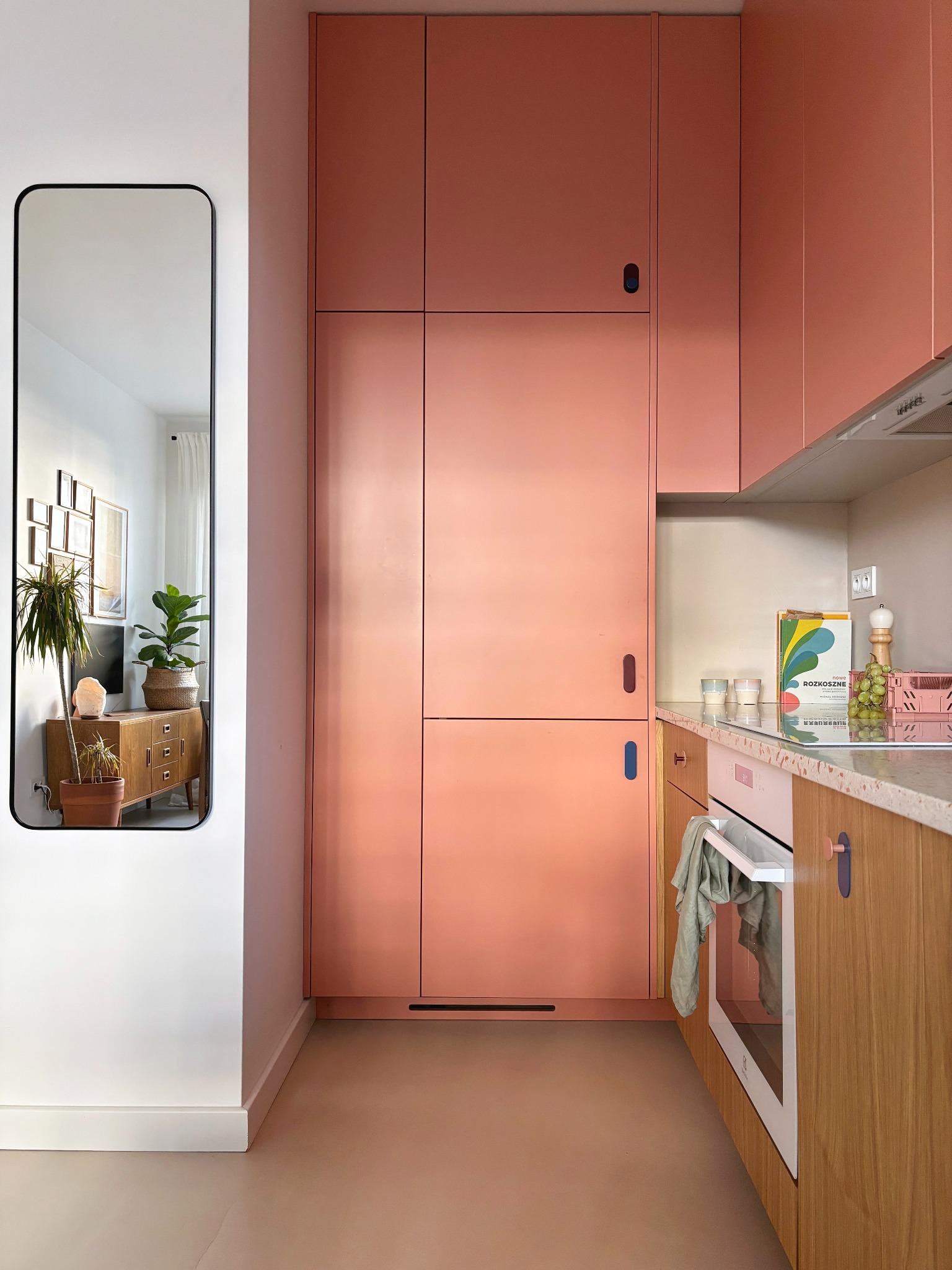 Wnętrze małej kuchni w mieszkaniu, zabudowa w kolorze łososiowym z piekarnikiem, blat z lastryko, lustro odbijające salon z roślinami.