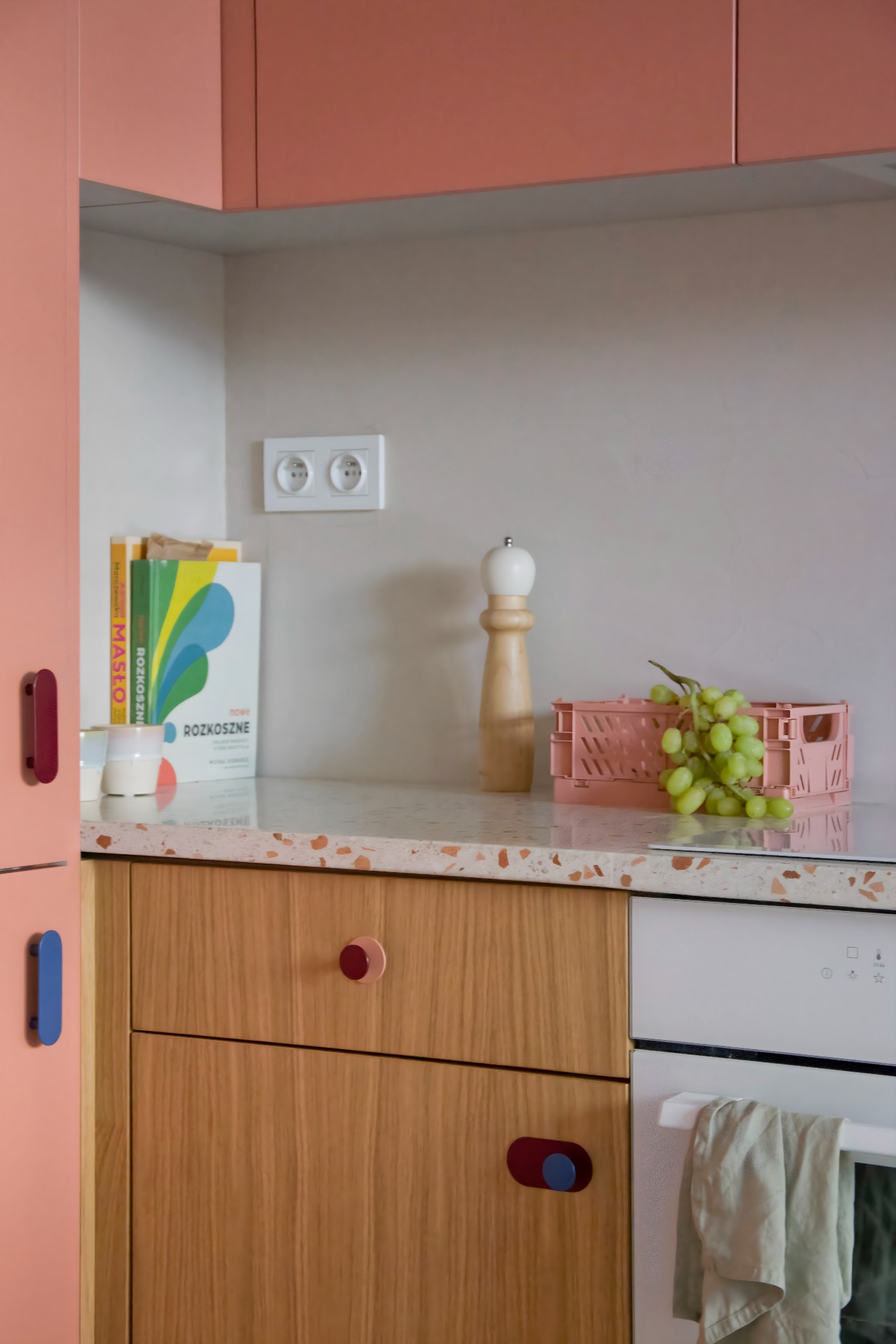Fragment kuchni z szafkami w odcieniach różu i drewna, blat z terazzo, drewniany młynek do pieprzu, różowa skrzynka z winogronami i książki z kolorowymi okładkami