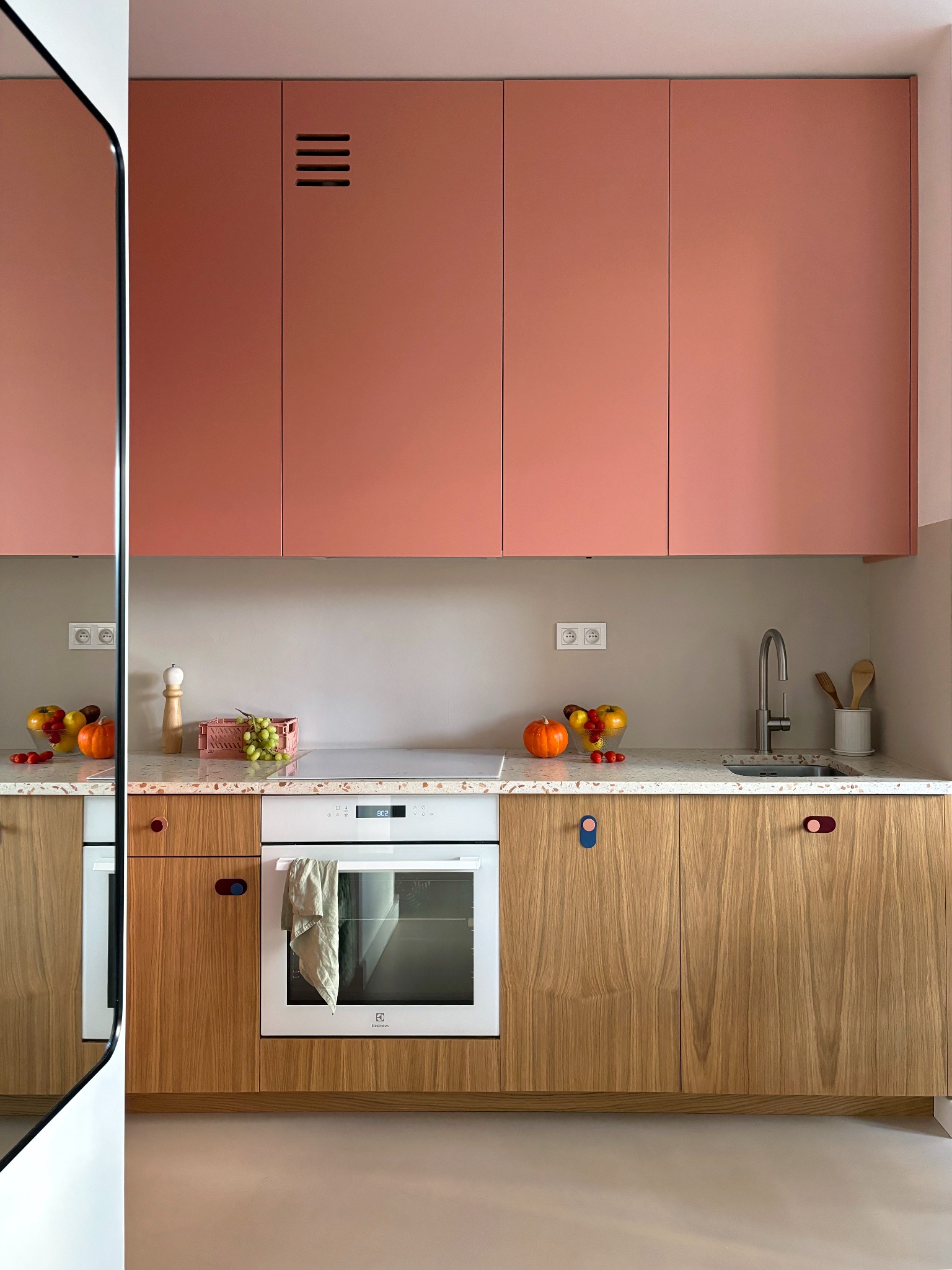 Nowoczesna kuchnia z drewnianymi szafkami dolnymi z nietypowymi uchwytami w kształcie pętli, koralowymi szafkami górnymi, blatem z lastryko i owocami na blacie.