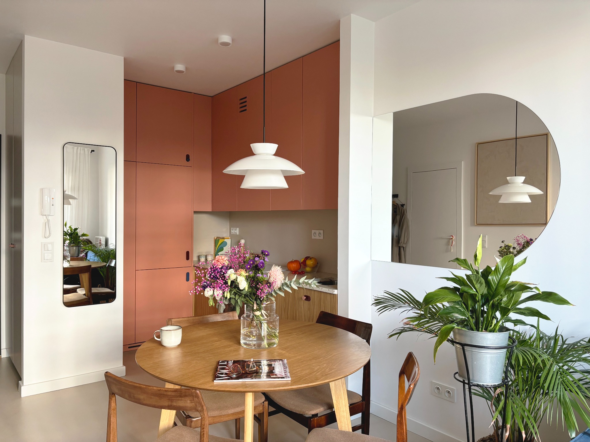 Jasne mieszkanie z okrągłym drewnianym stołem, bukietem kwiatów w szklanym wazonie, ceramicznym kubkiem i magazynem Vogue, z widokiem na różową kuchnię i rośliny doniczkowe.