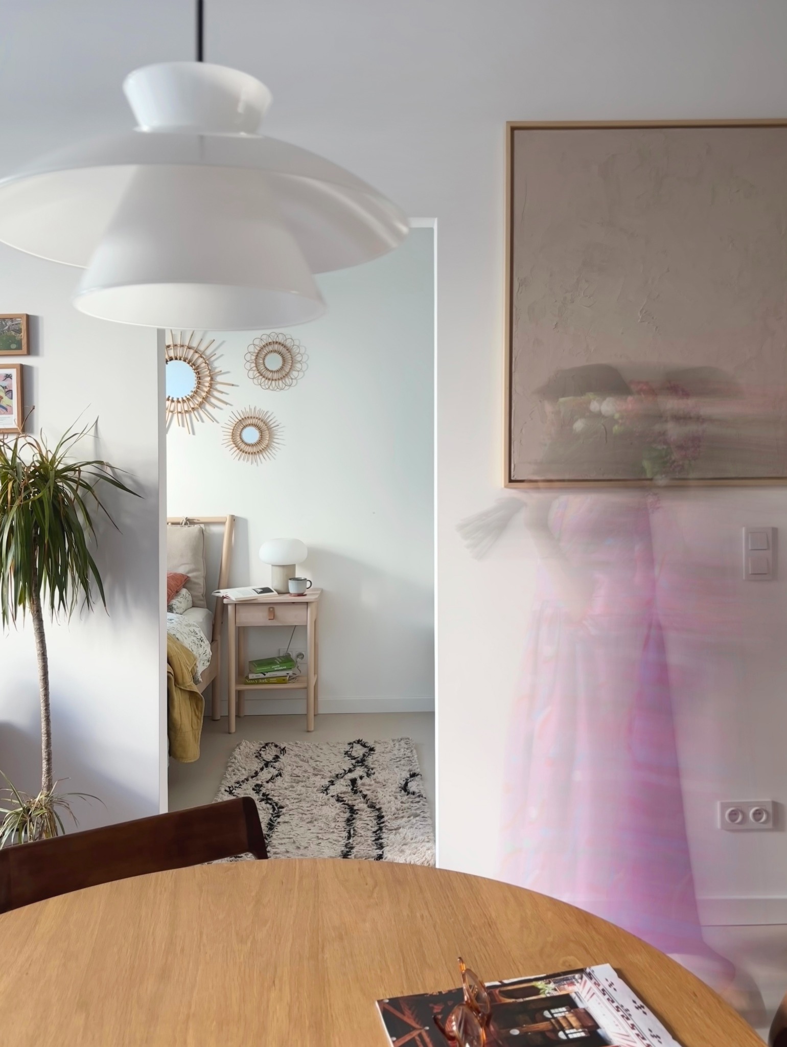 Wnętrze mieszkania z widokiem na sypialnię, okrągły drewniany stół z gazetą i okularami na pierwszym planie, duża biała lampa wisząca, roślina doniczkowa po lewej, na ścianie obraz i lustra...