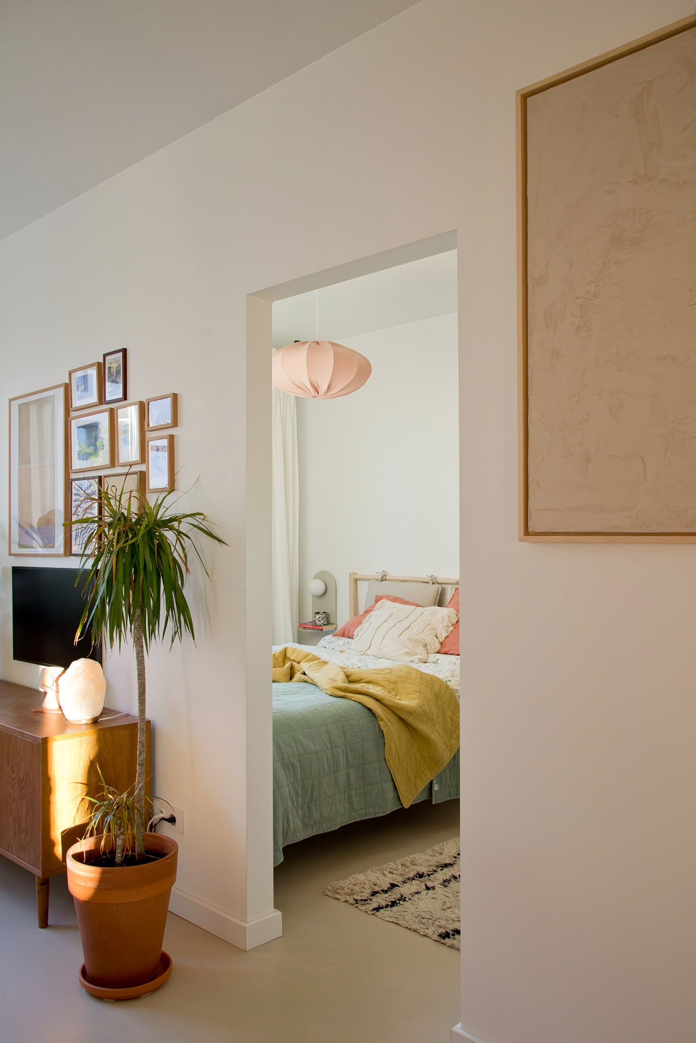 Widok na sypialnię z salonu, pastelowe kolory, łóżko z żółtą narzutą, lampa w kształcie kuli, galeria zdjęć na ścianie i roślina doniczkowa.