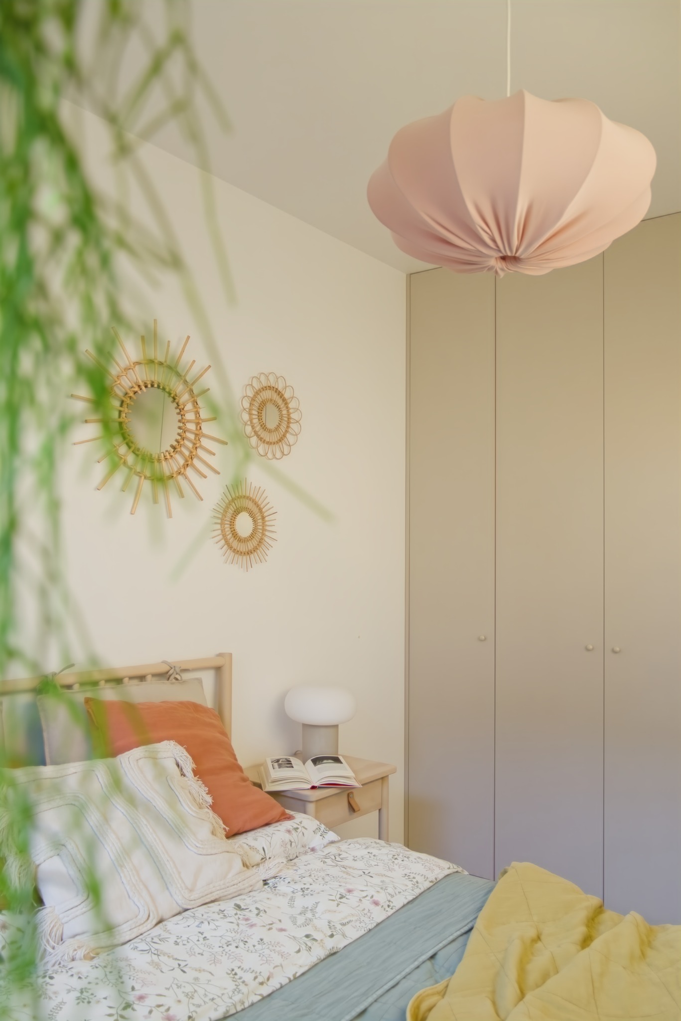 Sypialnia w pastelowych barwach z łóżkiem, szafą w zabudowie i okrągłymi lustrami na ścianie, oświetlona lampą w kształcie kwiatu.
