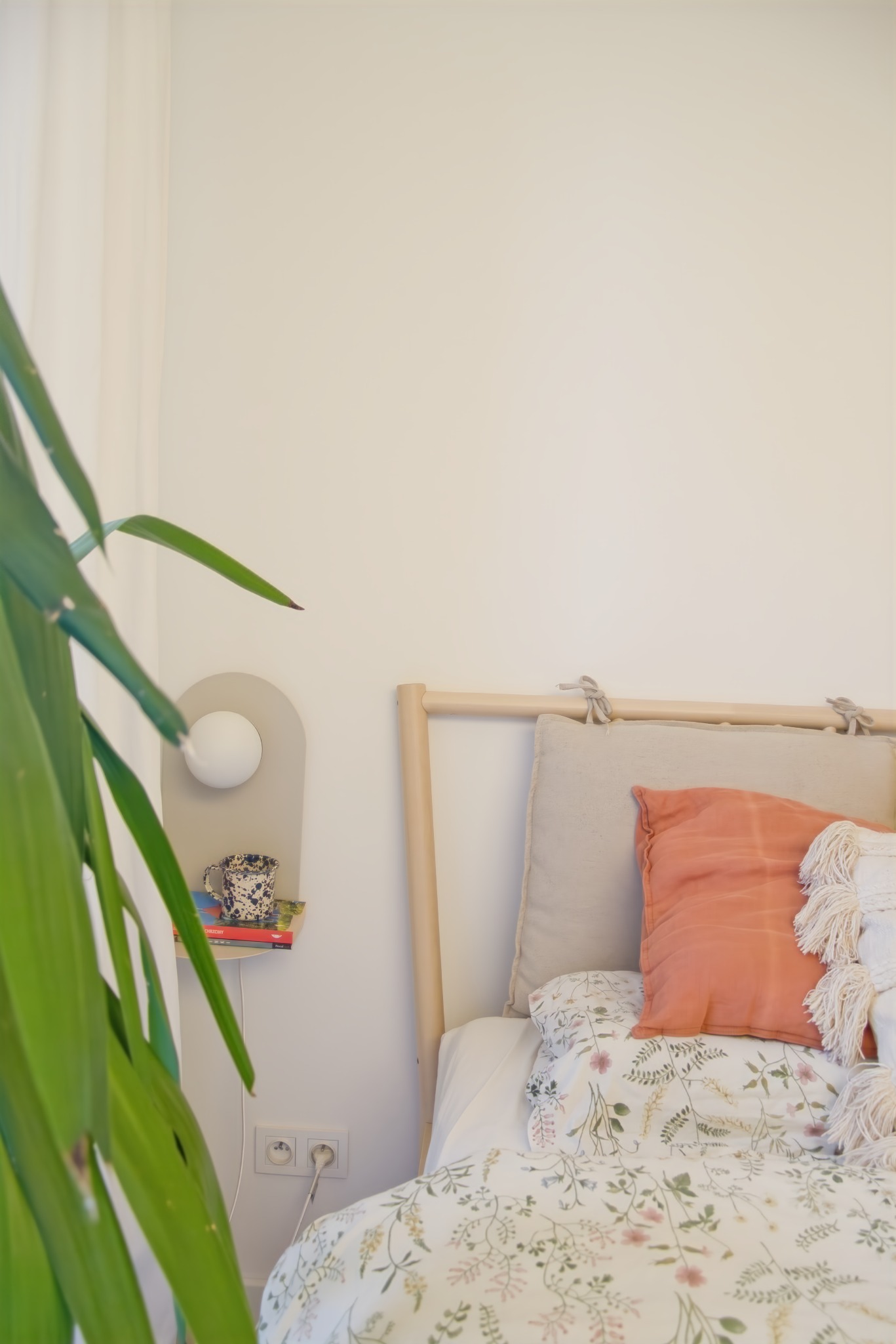 Jasna sypialnia w stylu skandynawskim z drewnianym łóżkiem, pościelą w kwiaty i pomarańczową poduszką, obok lampka nocna na półce z książkami i kubkiem, a w tle widoczna roślina doniczkowa.