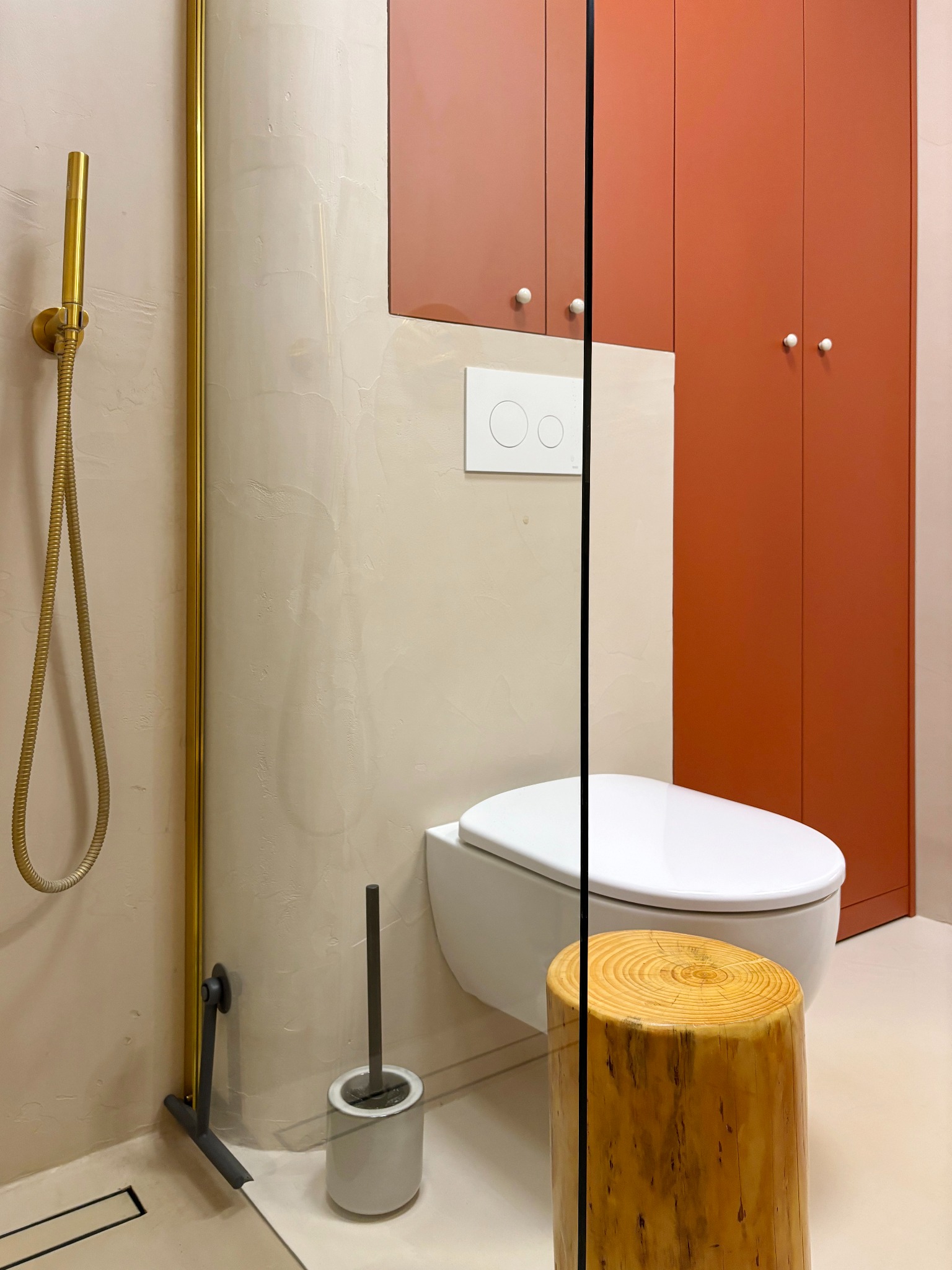 Nowoczesna łazienka z beżowymi ścianami, złotą armaturą prysznicową, podwieszaną toaletą i siedziskiem w formie pnia drzewa, uzupełniona o pomarańczowe szafki wiszące.