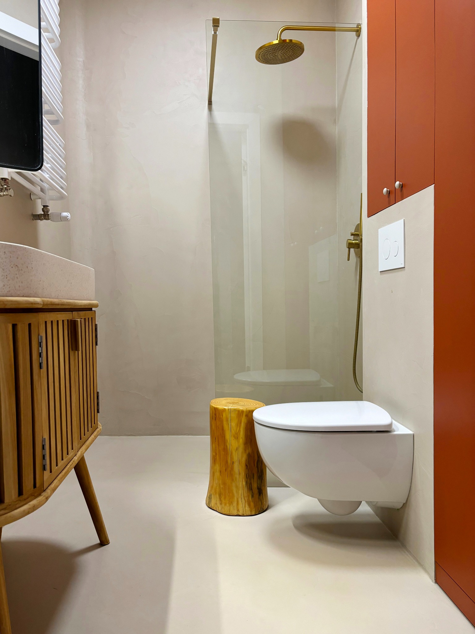 Nowoczesna łazienka z drewnianą szafką pod umywalką, kabiną prysznicową ze złotą armaturą i podwieszaną toaletą, uzupełniona o stołek imitujący pień drzewa.