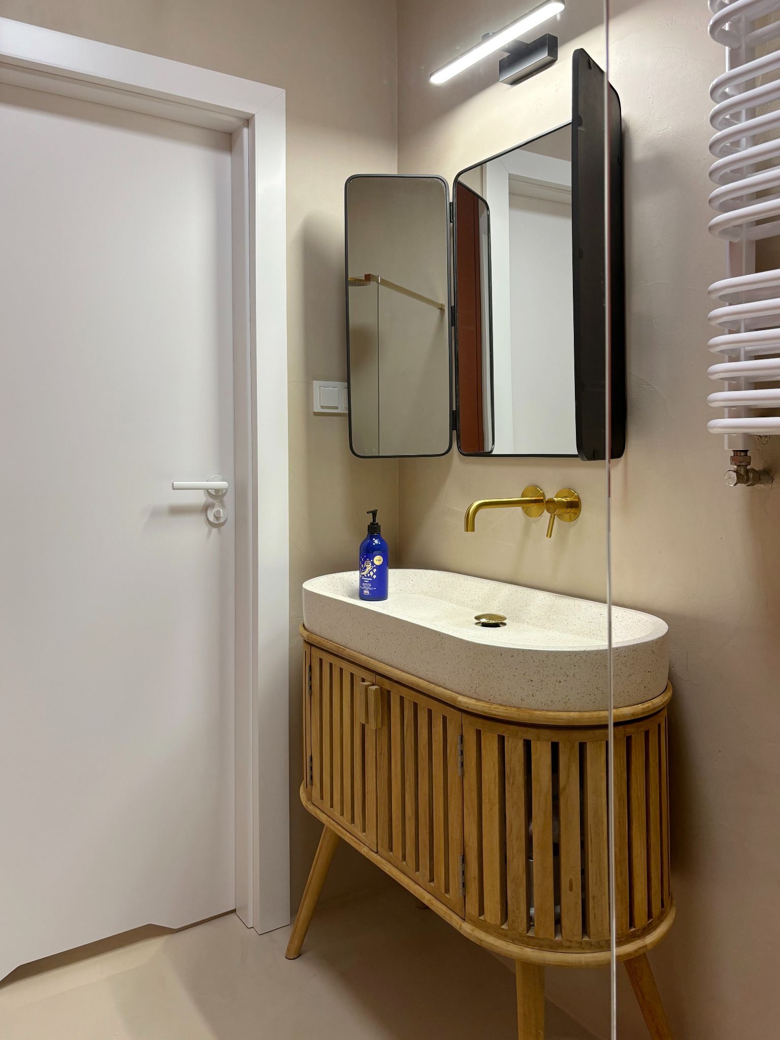 Nowoczesna łazienka z umywalką z terazzo na drewnianej szafce z lamelami, złotą armaturą i lustrem z otwieranymi skrzydłami.