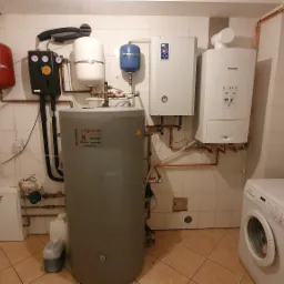 Kotłownia w piwnicy z szarym zasobnikiem ciepłej wody Viessmann, dwoma piecami, zbiornikami wyrównawczymi i pralką Bosch Navigator w rogu