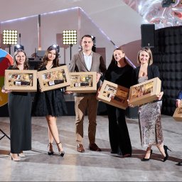 Paczka Events - Grupa osób w eleganckich strojach prezentuje upominki w kartonowych pudełkach z okienkami, widoczne koło fortuny z napisem 'paczka events' w tle, oświetlenie sceniczne.