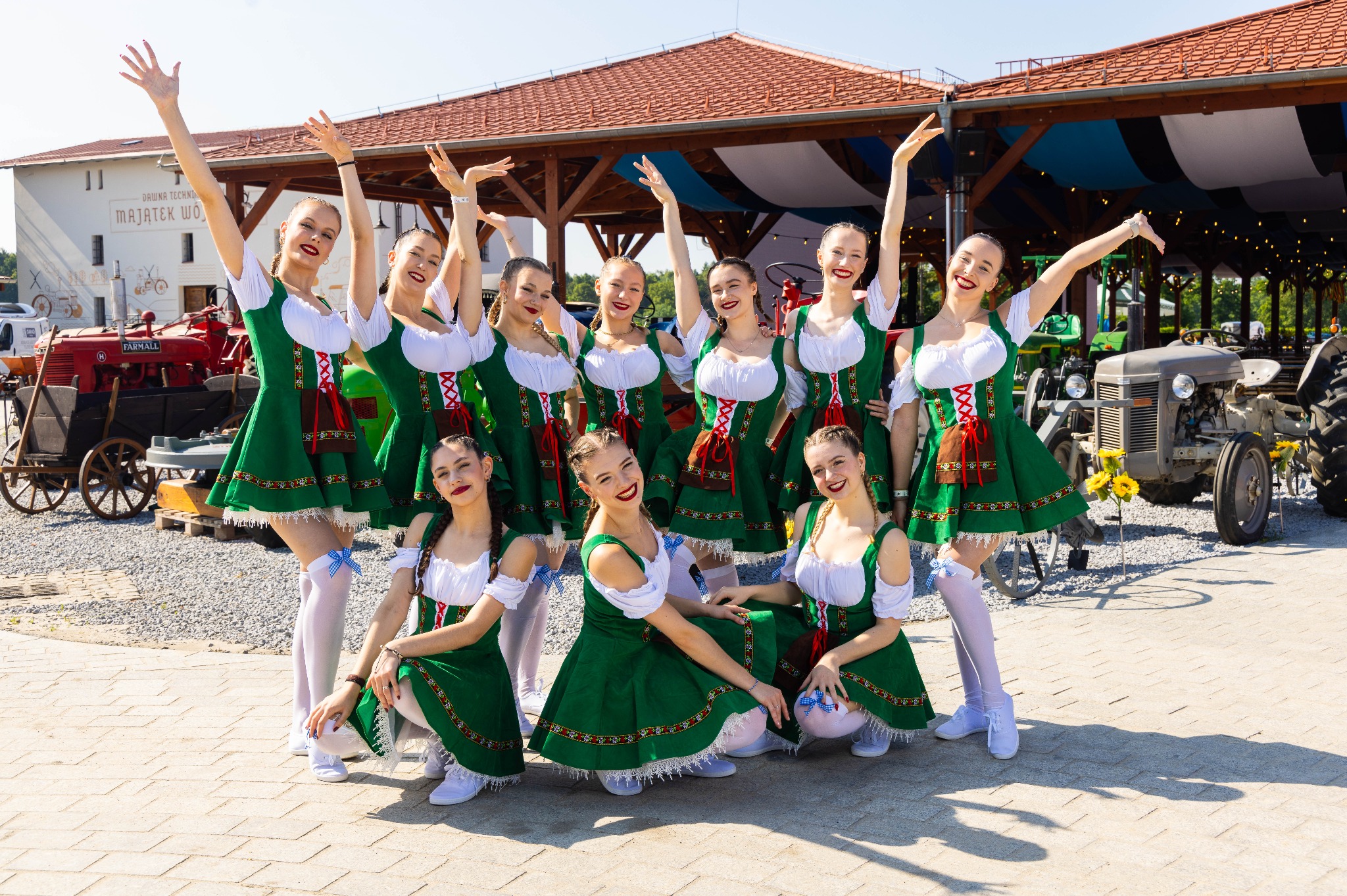 Grupa uśmiechniętych tancerek w tradycyjnych bawarskich strojach, pozujących na tle zabytkowych traktorów i budynku z muralem, na zewnątrz w słoneczny dzień.