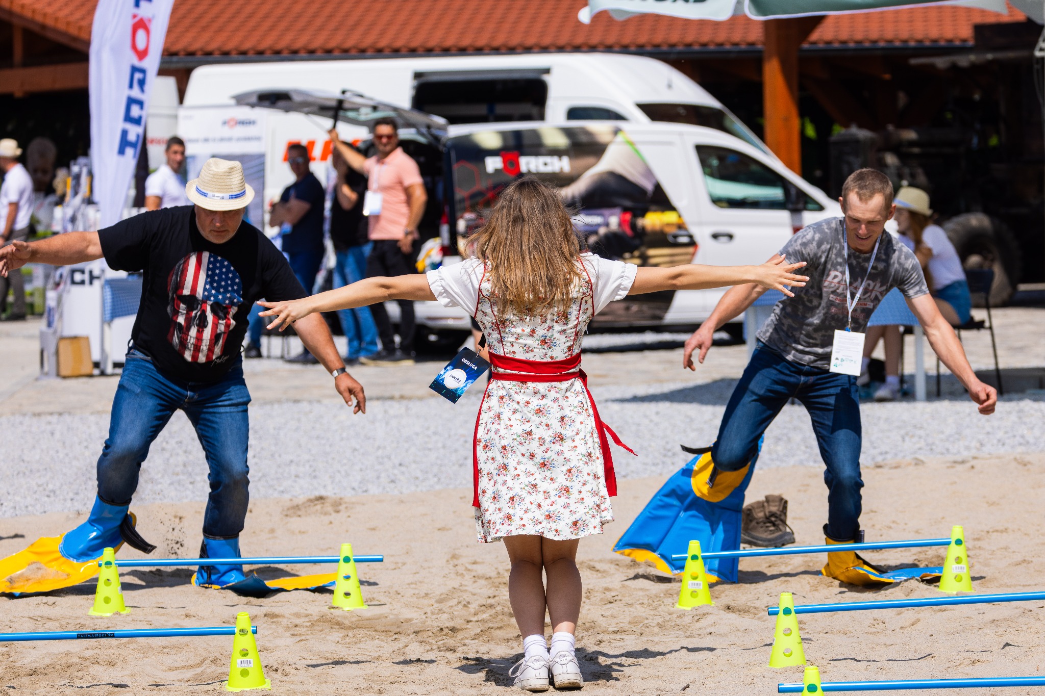 Uczestnicy w kaloszach na nartach, przechodzący przez tor przeszkód na piaszczystej plaży podczas imprezy integracyjnej, z widocznymi stożkami i barierkami.