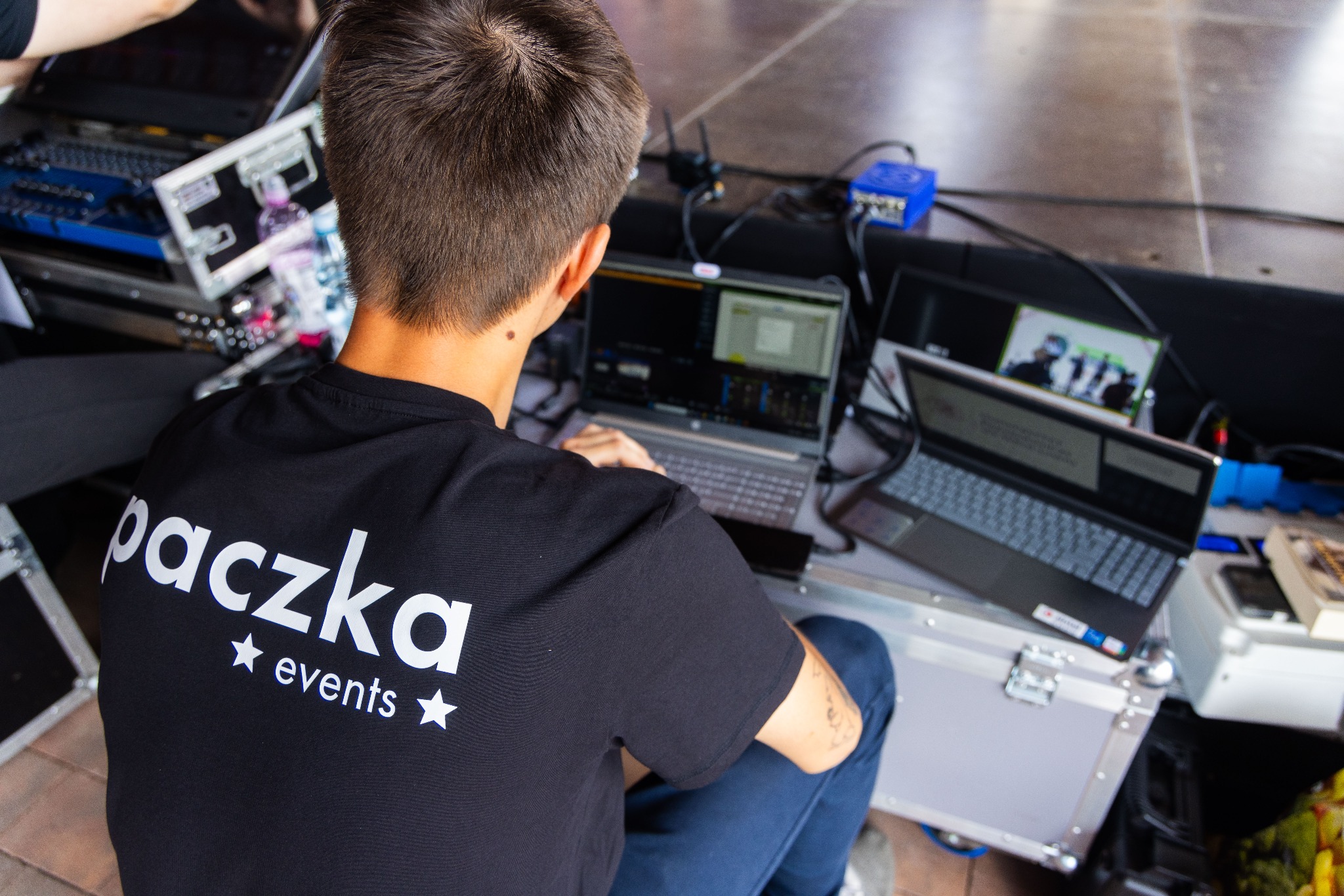 Osoba siedząca przed kilkoma laptopami, w koszulce z napisem 'paczka events', prawdopodobnie kontrolująca przebieg wydarzenia.