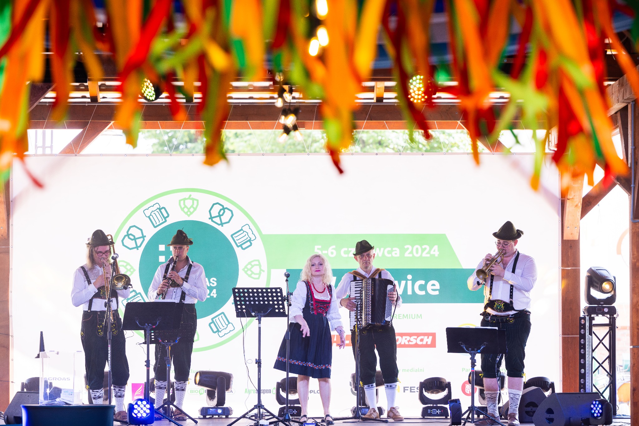 Zespół muzyczny w strojach bawarskich gra na scenie podczas wydarzenia plenerowego, z instrumentami dętymi, akordeonem i dekoracjami z kolorowych wstążek.
